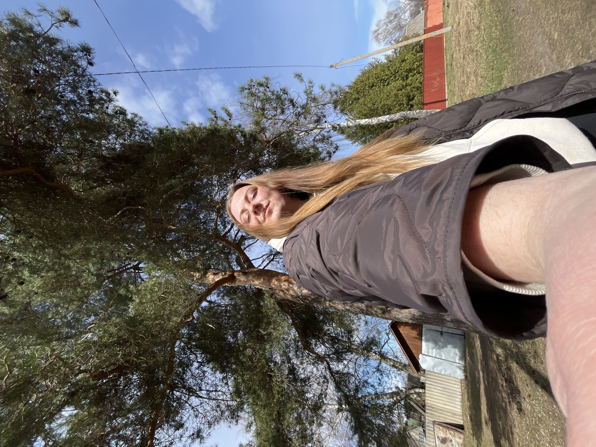A selfie shows a woman with long, blonde hair wearing a brown puffy jacket. Behind her is a large pine tree with a blue sky and scattered clouds visible through the branches. A small shed with a metal roof and a red fence are also in the background. Based on the location, the photo was taken in Раздолье, Russia.