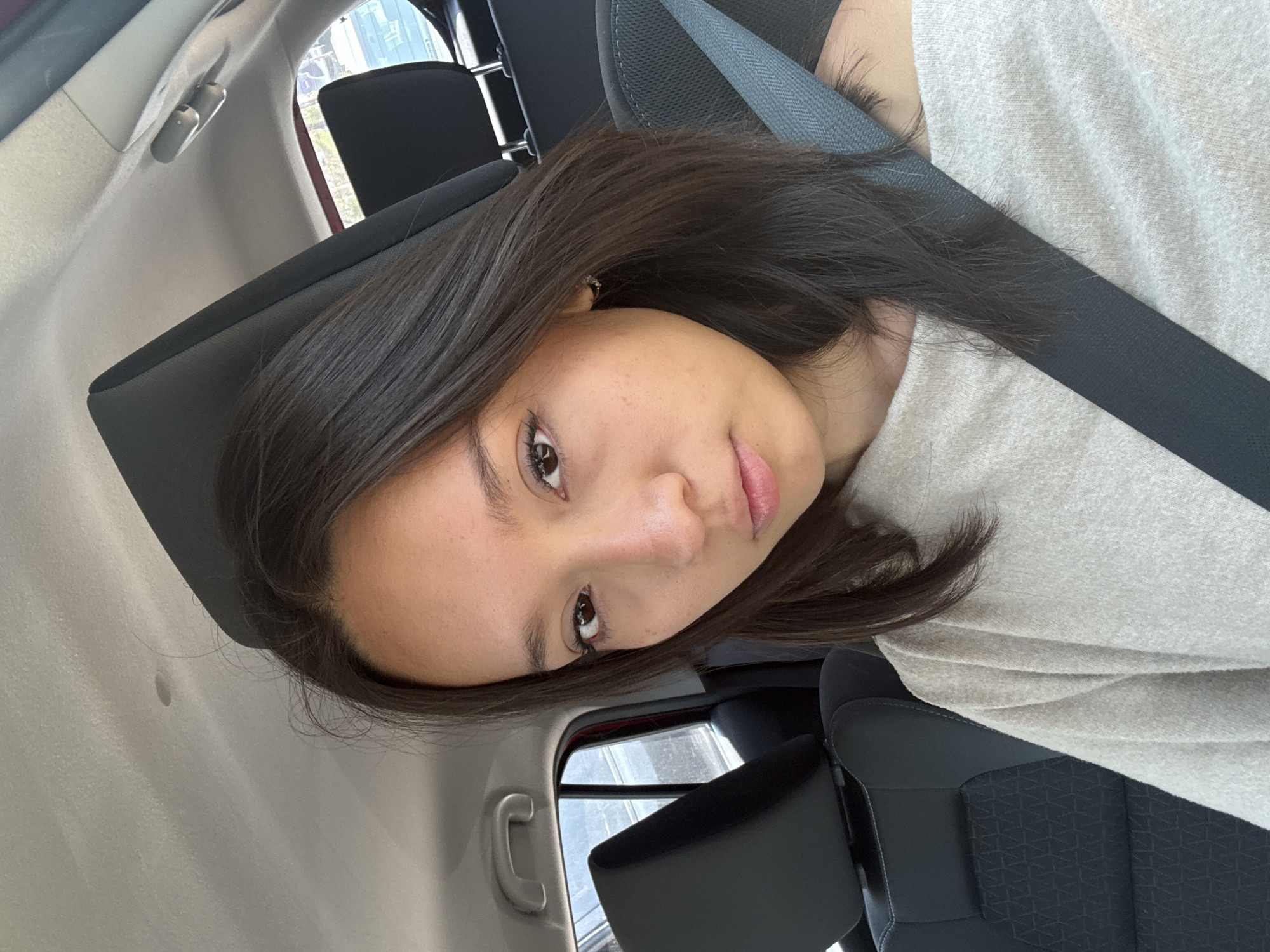 A young woman is captured in a close-up, slightly angled shot, appearing to be inside a car. Her dark, straight hair falls around her shoulders, and her gaze is directed towards the camera with a neutral to slightly contemplative expression. She has dark eyes, defined eyebrows, and full, naturally colored lips. She is wearing a light-colored, possibly grey or off-white, long-sleeved top. A dark seatbelt is prominently visible across her chest and shoulder, indicating she is seated in a vehicle.
The background reveals elements of a car's interior. A dark grey car seat, with a textured pattern on the lower section, is visible behind her. Bright daylight illuminates the scene, suggesting it is daytime, with glimpses of the outside visible through the car windows in the reflection. A car ceiling panel and a grab handle are also discernable. The overall atmosphere is casual and relaxed. No visible text appears in the image. This scene takes place in San Bernardino Tlaxcalancingo, Mexico.