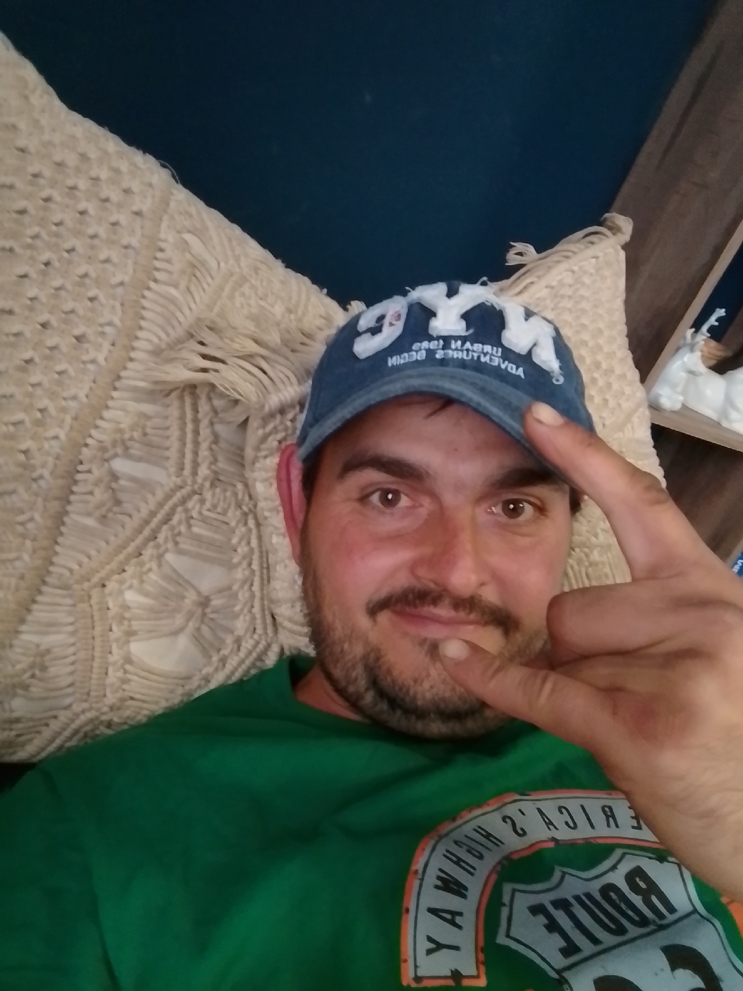 A Caucasian male, wearing a blue baseball cap with white lettering and a green t-shirt with a Route 66 design, is shown from the chest up, lying down. He is holding up his right hand with his index and pinky fingers extended in a rock-on gesture. He is looking at the camera with brown eyes and has a mustache and beard. Behind him, a cream-colored macrame wall hanging and a dark blue wall are visible. On a wooden shelf to the right, a white deer figurine is present. The location is not specified as Wedel, Germany.
