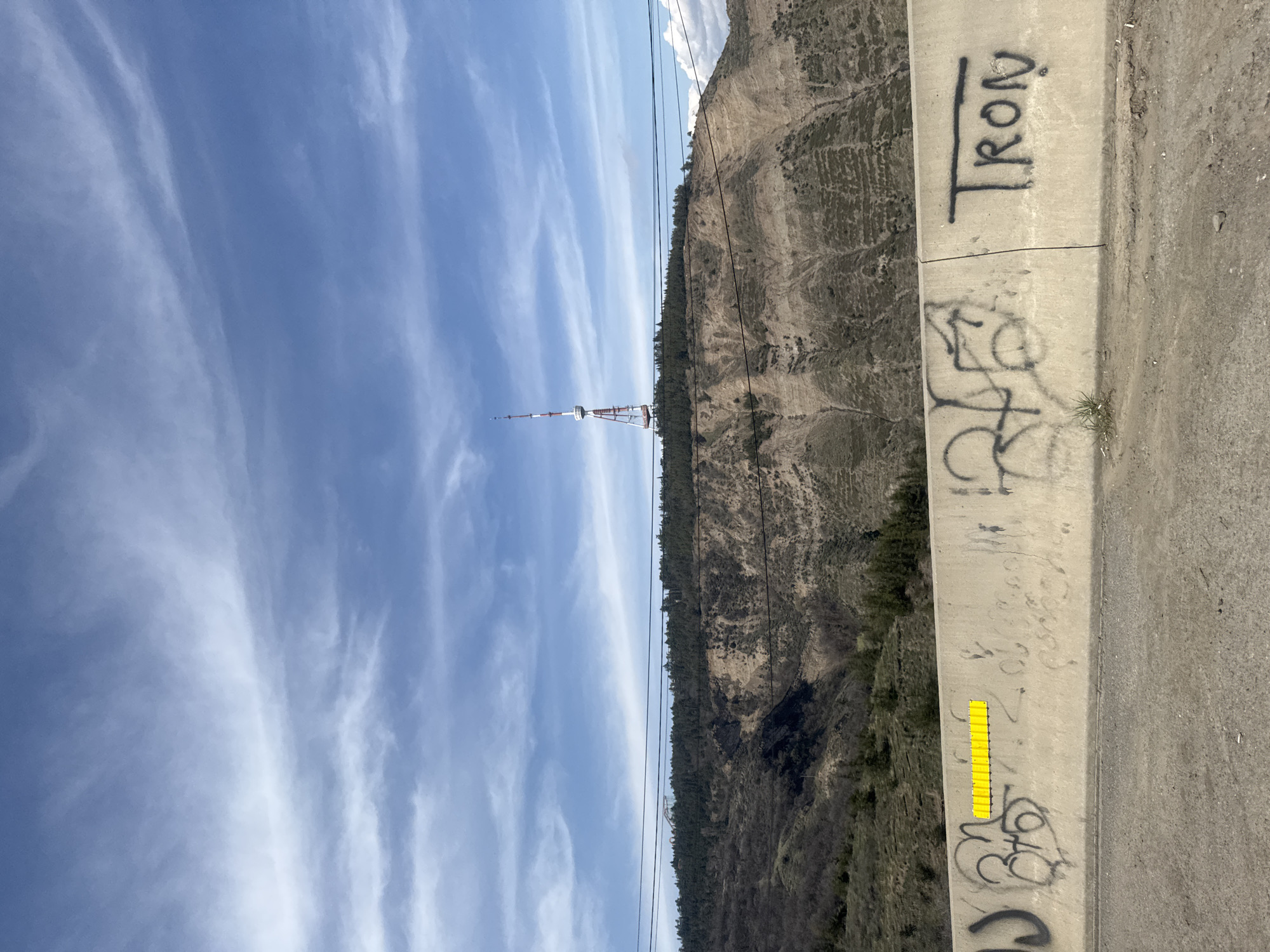 The image shows a view from a high vantage point in Tbilisi, Georgia. In the background, there is a tall communication tower situated on a hilltop. The sky is blue with streaky clouds. A hillside covered with vegetation rises to the right of the tower. In the foreground is a concrete barrier. Graffiti is on the barrier. A yellow, rectangular sticker is also attached to the barrier.