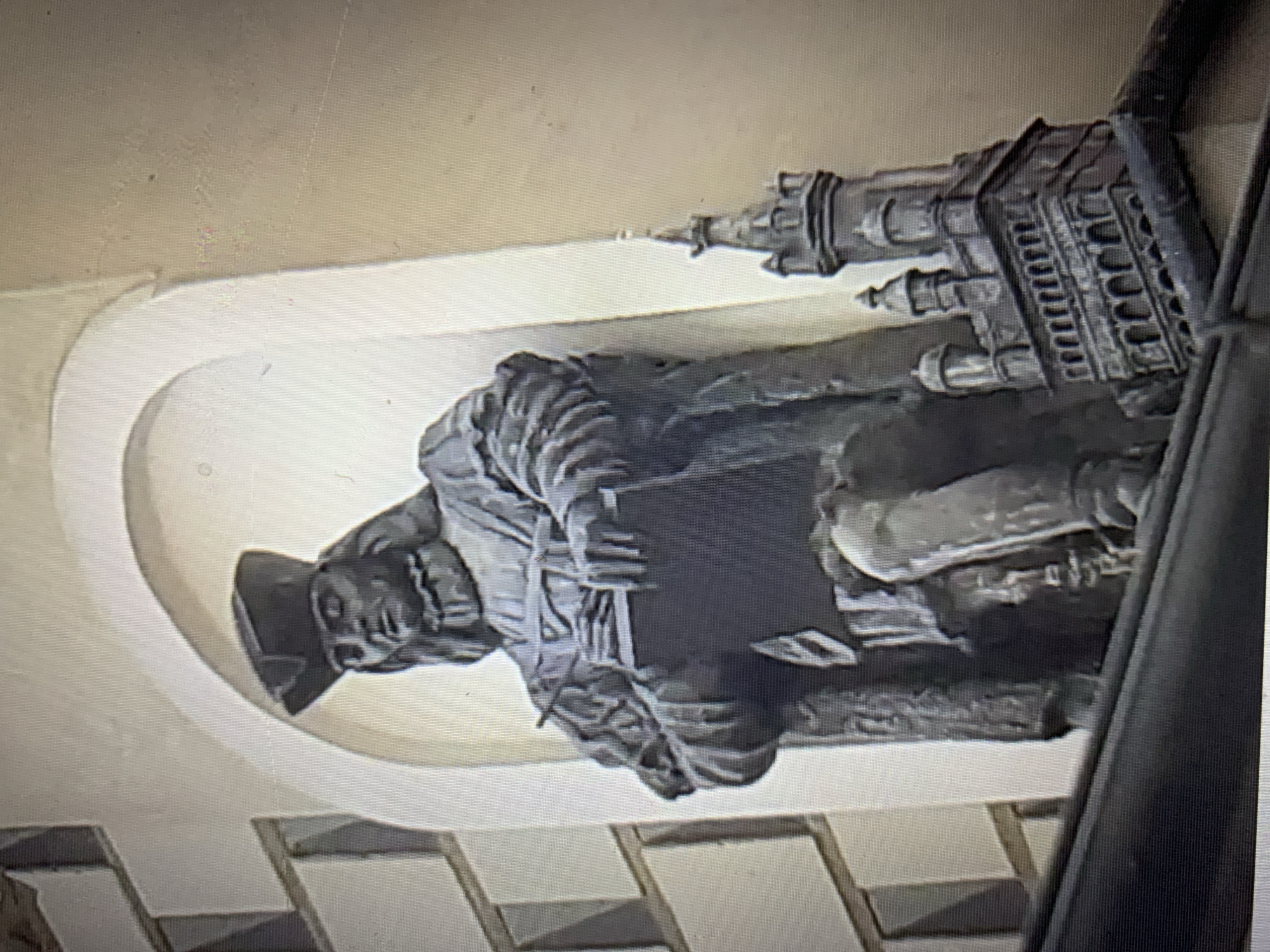 A dark, low-resolution image shows a stone statue of a man wearing a hat and holding a book. The statue is recessed into an archway on a building. To the right of the statue is a smaller stone structure resembling a castle. The background wall of the building appears to be beige. The image quality is poor, with visible pixelation and some color distortion. The location in the city of Lymanka, Ukraine, is not discernible from the image itself.