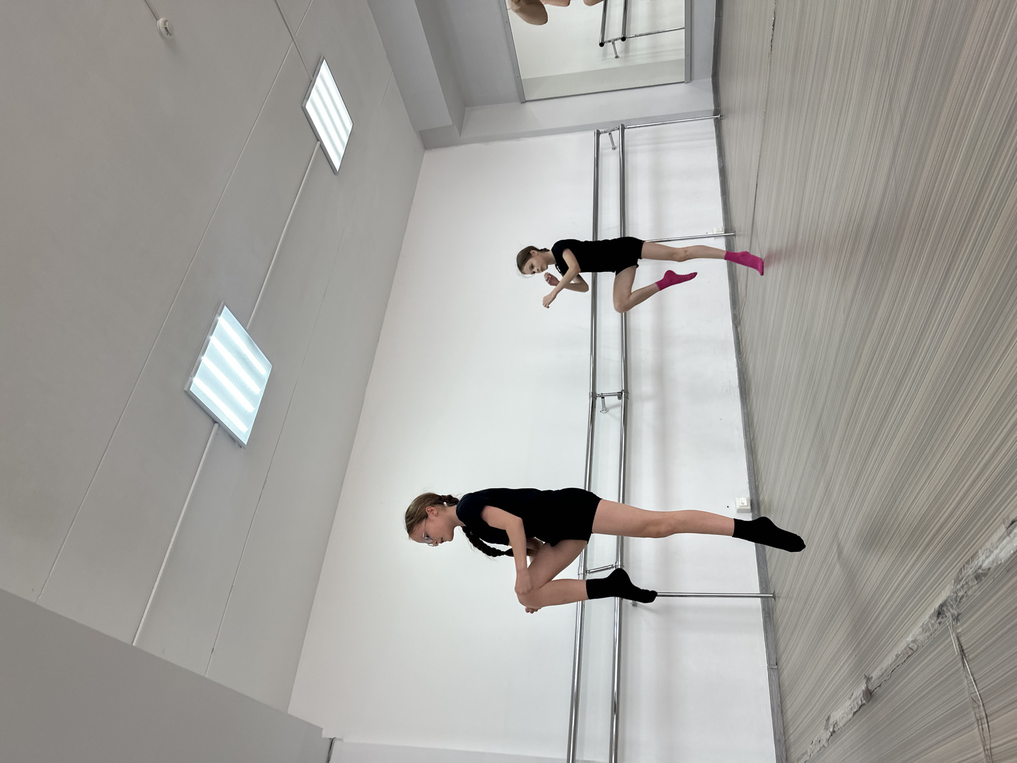 Two young girls in black dance attire are practicing in a dance studio. The studio has white walls, a barre, and large mirrored walls. In the foreground, a girl with blonde hair in a braid is positioned on the barre, with her right leg extended and bent at the knee, and her left leg also bent. She is wearing black socks. Behind her, another girl, also with blonde hair and a braid, is in a similar position on the barre, with her right leg extended and bent, and her left leg extended behind her. She is wearing pink socks. Both girls appear to be focused on their movements. The lighting in the studio is bright, coming from ceiling lights. The overall scene suggests a dedicated practice session. The location is Votkinsk, Russia.