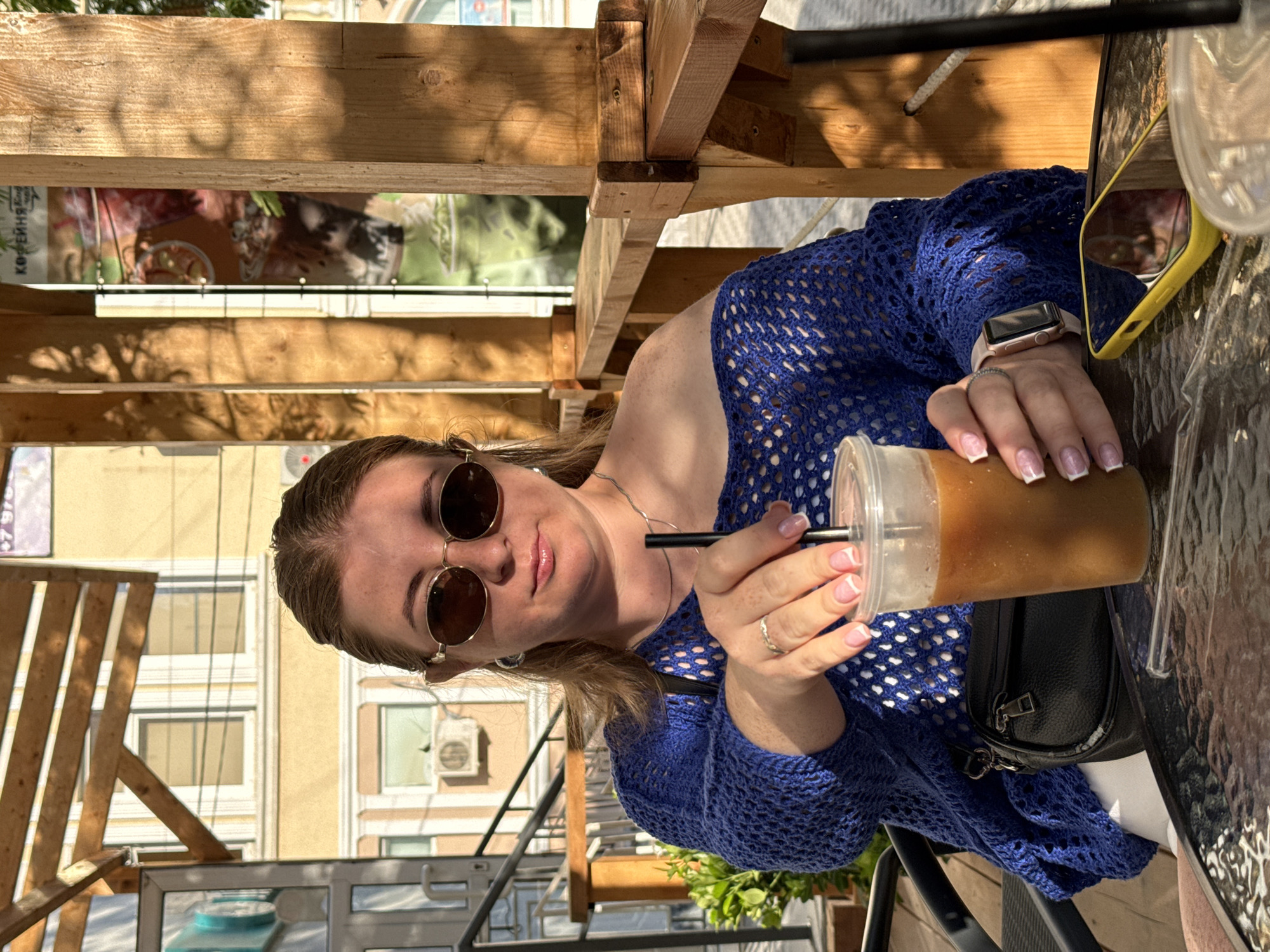 A woman with blonde hair, wearing sunglasses, a blue knitted off-the-shoulder top, and a white shirt, sits at a table outdoors, holding a plastic cup of iced coffee. She is wearing a ring on her left hand and a smartwatch on her right wrist. A black fanny pack is slung across her shoulder. A yellow smartphone is visible on the table next to the coffee cup. The background shows a building with cream-colored walls and windows, and wooden structures that appear to be part of an outdoor seating area. The lighting suggests it is daytime. The scene takes place in an urban setting, possibly a cafe or restaurant.