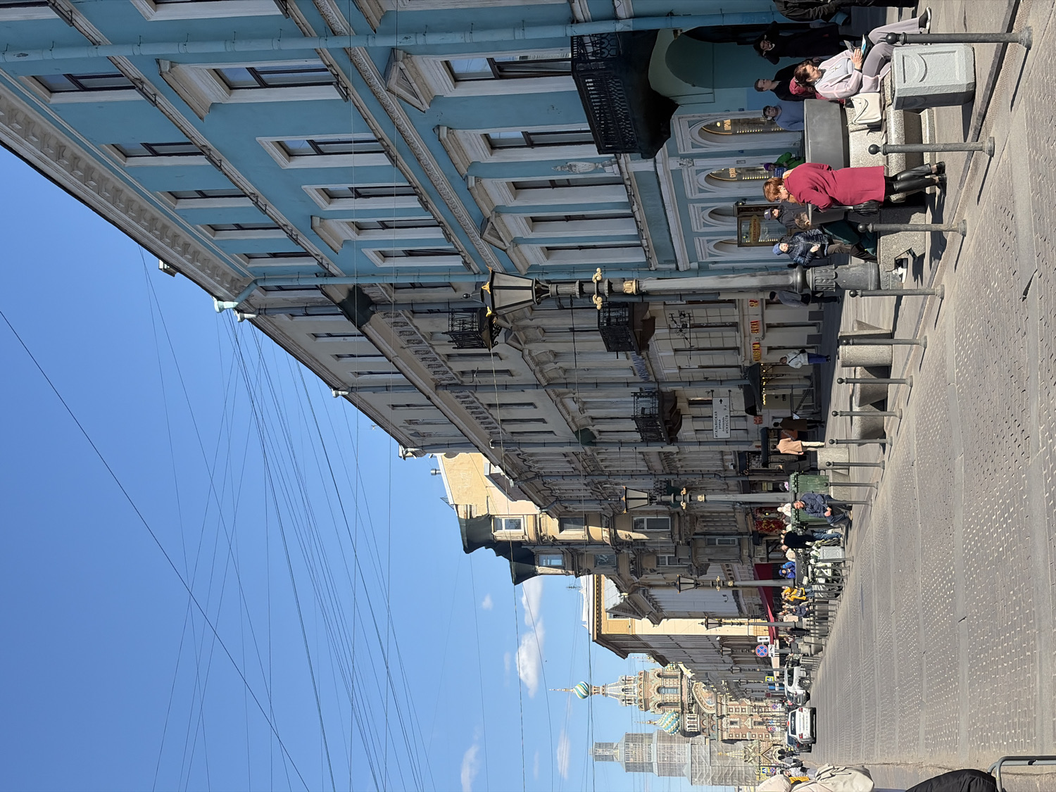 This image captures a daytime street scene in Saint Petersburg, Russia, under a clear, bright blue sky with sparse clouds. Historic, ornate buildings line the right side of the street, extending into the distance. The closest building features a light blue facade with white architectural trim and numerous windows. A complex network of thin overhead wires crisscrosses the sky above the street.
Several pedestrians are visible on the sidewalk to the right. In the mid-ground, a woman in a red coat stands near a series of decorative bollards. Other individuals are seen walking, standing, and sitting on low stone structures along the pavement. Further down the street, cars are sparsely parked or moving.
A prominent landmark, the multi-domed Church of the Savior on Spilled Blood, with its distinctive colorful onion domes, is clearly visible in the background, confirming the location. The scene suggests a typical, sunny day in the city, with people engaged in daily activities. Visible Cyrillic text on various shop signs includes "СУШИ" (SUSHI) and partially "ВЫВЕСКА" (VЫVESKA - signboard/billboard).