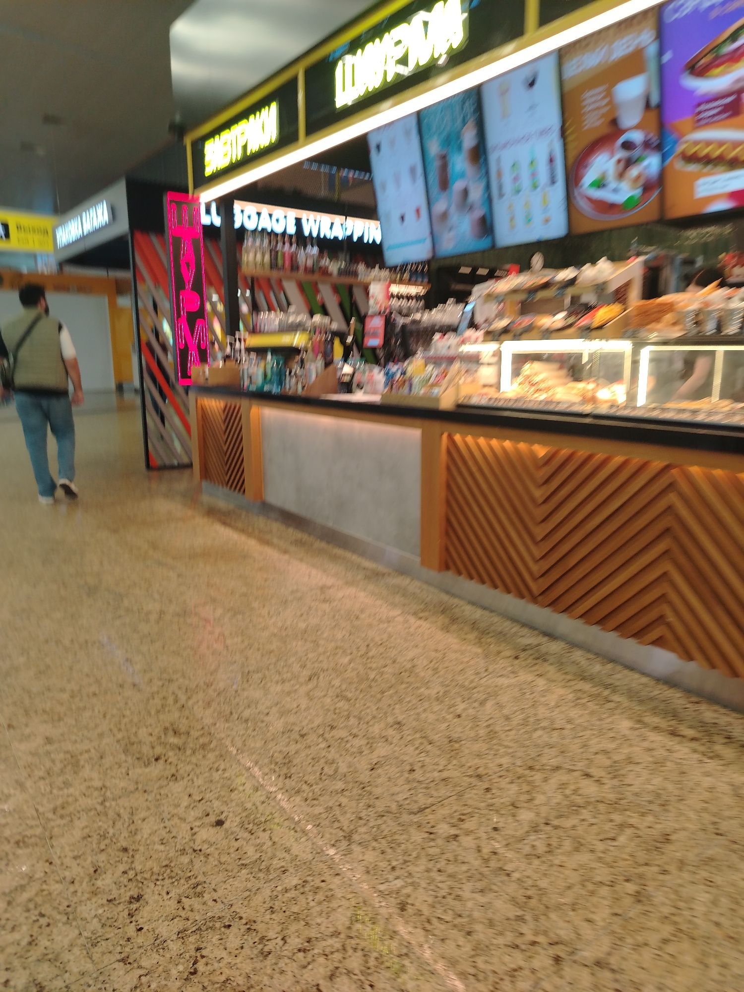 The image shows a food counter in a public area. The counter has a wooden front with a zigzag pattern and a gray central section. There's a lighted display case on top of the counter showing baked goods. Above the counter is a display of beverages and a menu. A red neon sign is present near the left edge of the counter. A person in a vest and jeans is walking away from the camera on the left side of the image. A sign in Russian is partially visible above the person. The location is Khimki, Russia, as suggested by the Russian text on the signs. The floor is a patterned tan color. The scene appears to be inside an airport or a shopping mall.