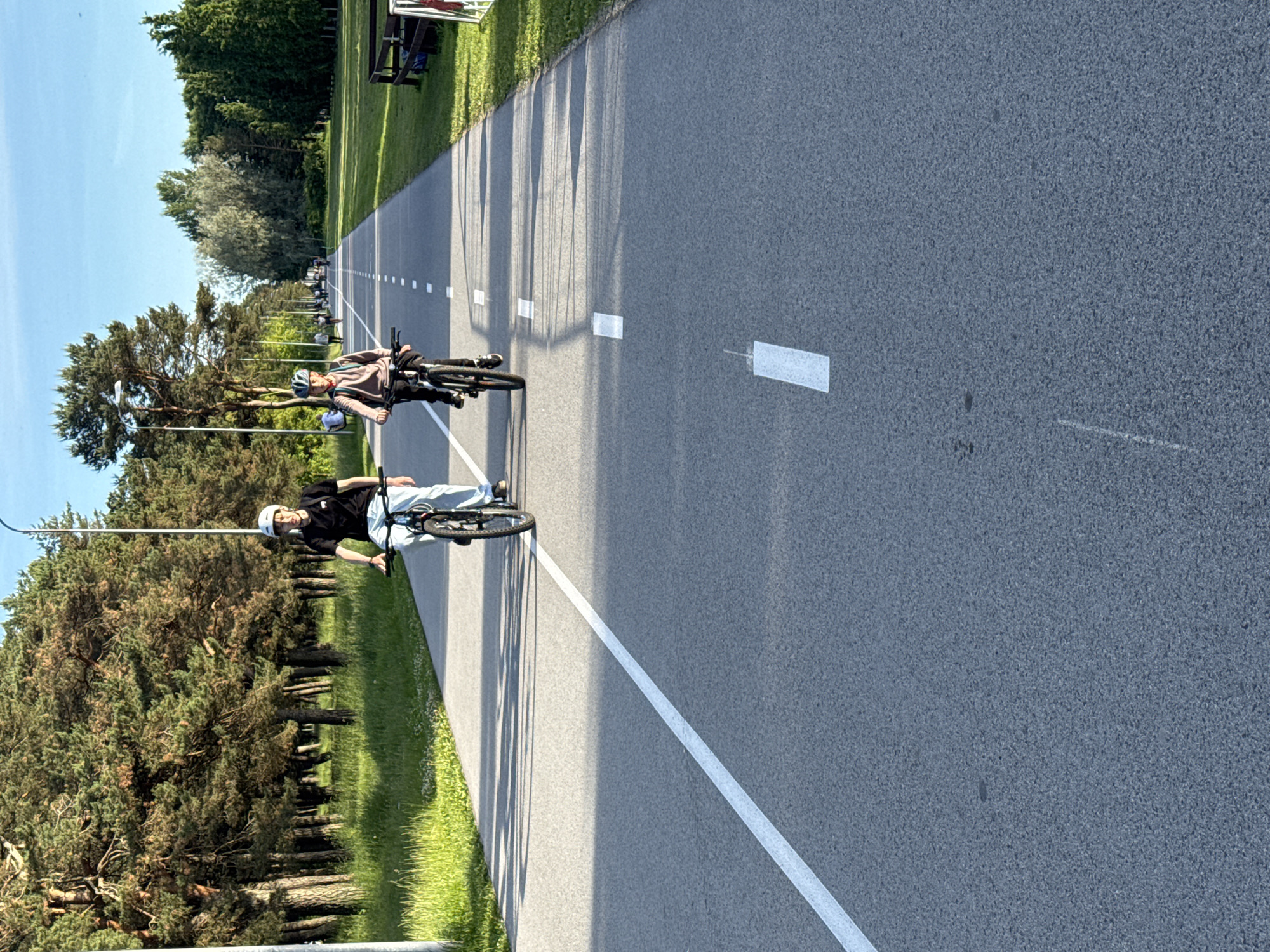 Two individuals, appearing to be children or teenagers, are cycling on a paved bike path. The person on the left is wearing a black t-shirt, light-colored pants, and a white helmet. They are riding a bicycle with a light-colored frame. The person on the right is wearing a light brown long-sleeved shirt, dark pants, and a helmet. They are riding a bicycle with a dark frame. Both individuals are facing forward, cycling away from the camera. The bike path is asphalt with white lane markings. To the left of the path is a grassy area with trees, including pine trees. To the right of the path is a grassy area with more trees and park benches. The sky is clear and blue. The scene appears to be a park or recreational area in the city of Tallinn, Estonia. The lighting suggests it is daytime, likely late afternoon or early evening due to the long shadows.