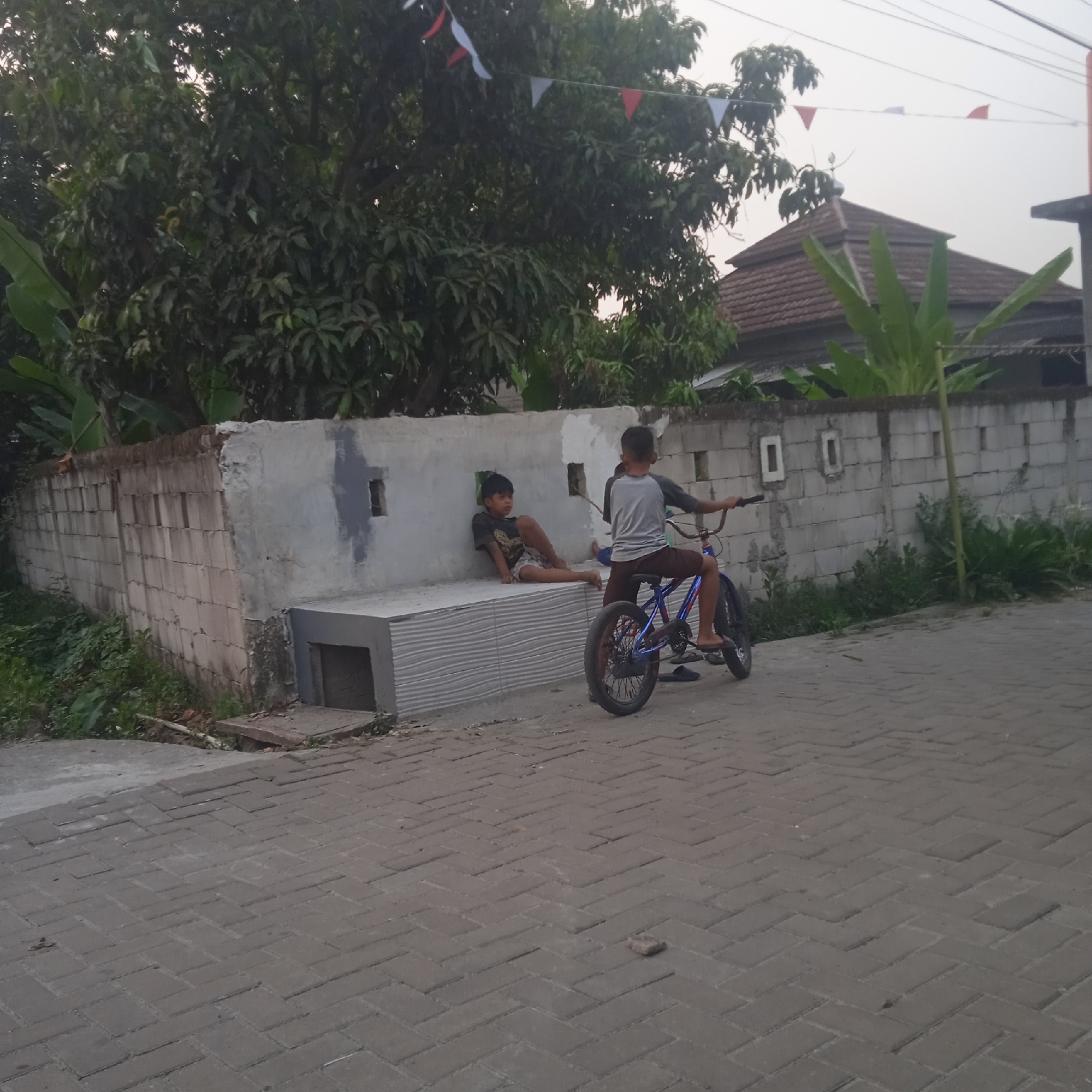 In Situterate, Indonesia, two young boys are present in an outdoor environment. One boy is seated on a low concrete structure, leaning against a partially repaired wall made of concrete blocks. He is wearing a dark t-shirt and light-colored shorts. The other boy is on a bicycle with blue accents, wearing a grey t-shirt and dark shorts. He is holding onto the handlebars of the bicycle. The bicycle has a metal frame and appears to be a BMX-style bike. The background shows a wall of concrete blocks, some overgrown vegetation, and trees. Overhead, a string of red and white triangular flags is visible, suggesting a celebration or decoration. The ground is paved with interlocking concrete bricks laid in a herringbone pattern. The overall lighting suggests it is either early morning or late afternoon.