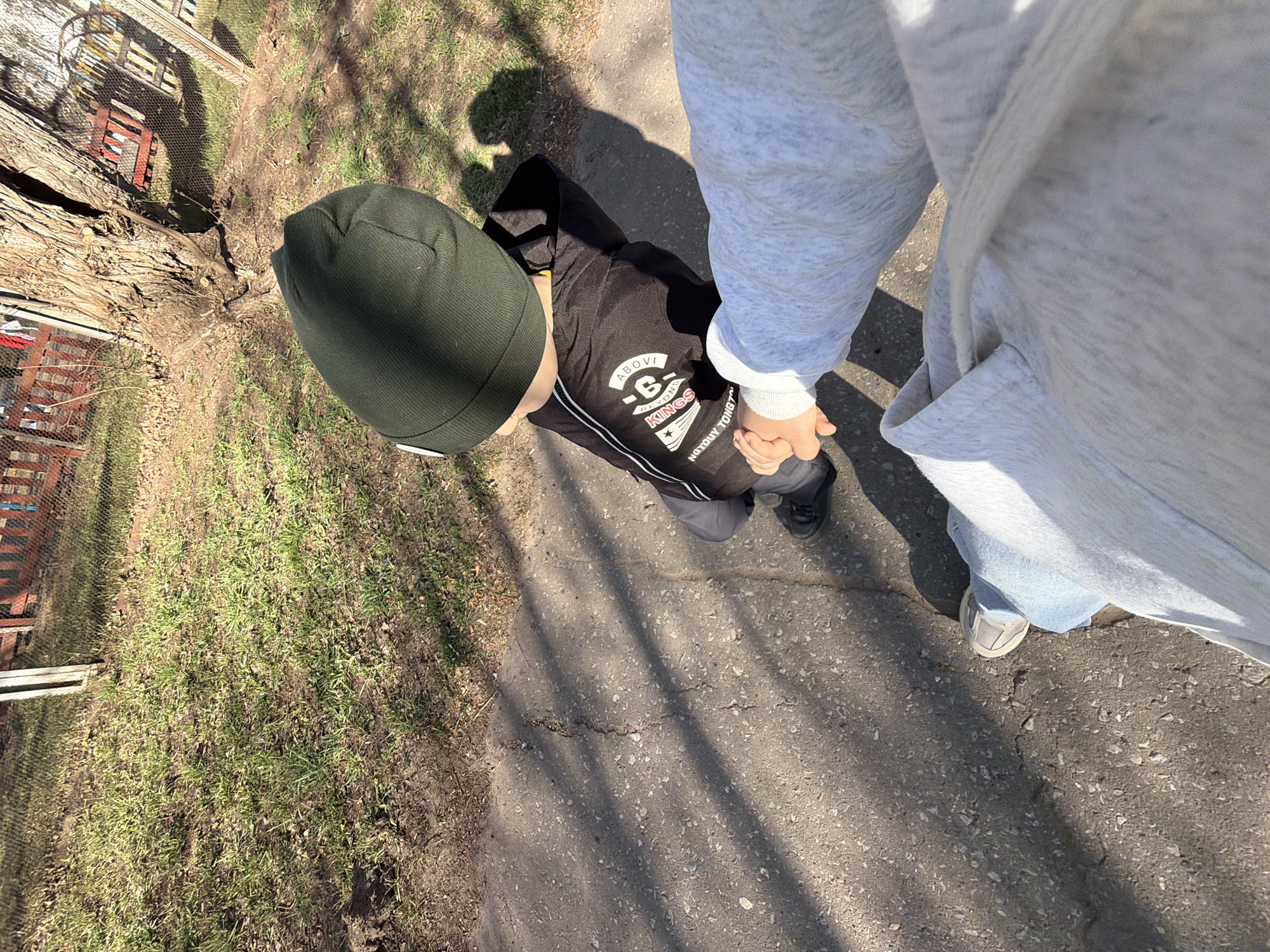 A photograph taken outdoors shows a child and an adult holding hands while walking on an asphalt path. The child is wearing a dark green beanie, a black jacket with white accents, and gray pants with black shoes. The adult is wearing a light gray hoodie, blue jeans, and gray sneakers. The path is cracked and uneven, with patches of grass and dirt on either side. A wooden fence and trees are visible in the background. The location is likely a park or residential area in Chaikovsky, Russia, based on the visible Russian text on the child's jacket. The photo appears to be taken from an overhead perspective.