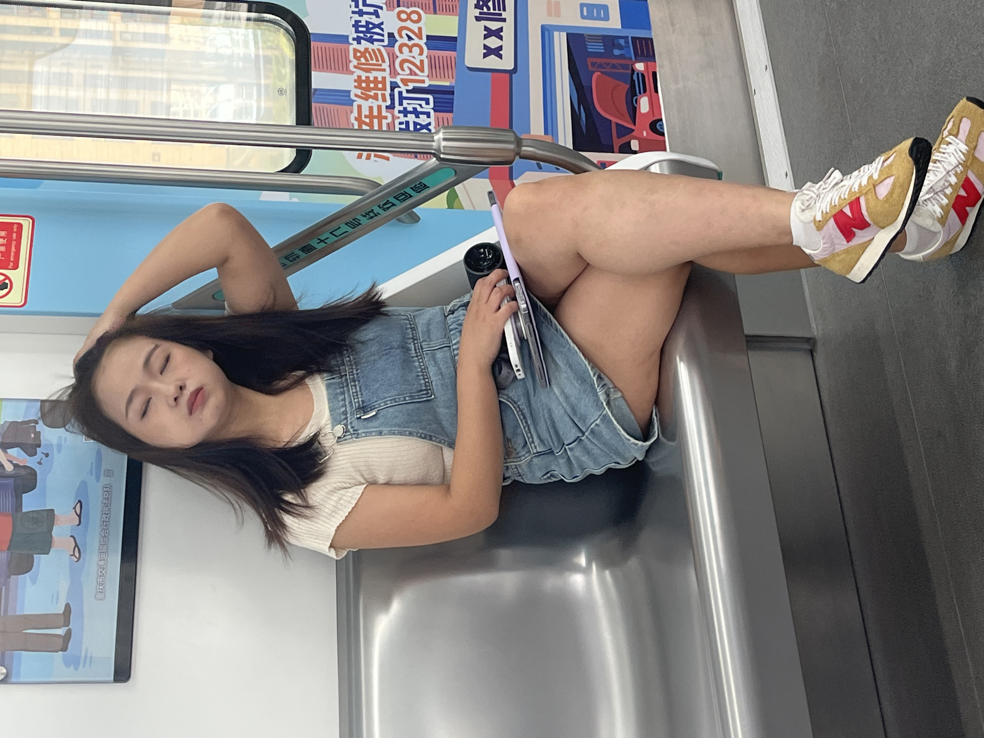A young East Asian woman with dark hair is sitting on a metallic subway seat, leaning back with her eyes closed and one hand resting on her head. She is wearing a denim overall dress over a white t-shirt and a pair of colorful athletic shoes. She is holding a tablet or similar electronic device in her lap. The subway car interior is visible, with metallic walls and seats, and windows showing a blurred city scene outside. There are Chinese characters on a poster on the wall behind her. The overall atmosphere suggests a moment of rest or sleep during a subway commute. The location is identified as the city of Tiaodeng, China.