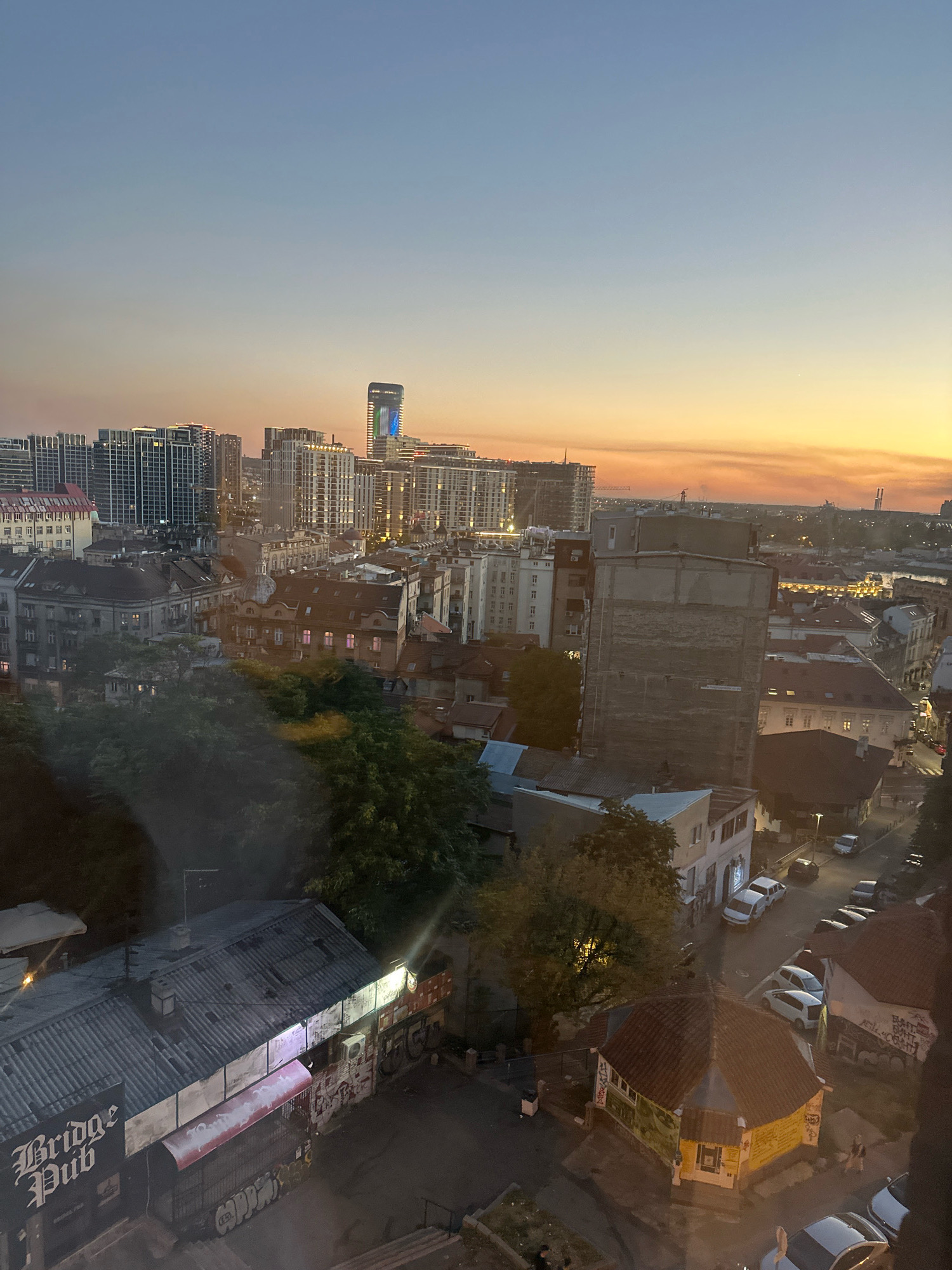 An aerial perspective captures an urban cityscape at dusk, likely in Belgrade, Serbia, as suggested by the architectural style and the presence of the "Bridge Pub." The sky transitions from a pale blue to a warm orange hue near the horizon, indicating the setting sun. In the background, modern high-rise buildings with illuminated windows dominate the skyline, contrasting with the older, lower-rise structures in the mid-ground and foreground.
The mid-ground features a dense arrangement of buildings with pitched roofs, many of which show signs of age. Some windows emit a soft, colored light, suggesting habitation or interior lighting. A prominent, taller building with a stark, unfinished facade stands out in this section. Green foliage from trees is visible, breaking up the urban density in several areas, particularly in the mid-left of the image.
The foreground is dominated by a lower building with a corrugated metal roof, identified by a sign that reads "Bridge Pub." This establishment appears to be a commercial space, with graffiti visible on its walls and awnings. Adjacent to the pub, a small, hexagonal building with a colorful, graffiti-covered exterior is present. The street scene reveals several parked cars, including white sedans and other vehicles, along with a few indistinct human figures walking on sidewalks or in the vicinity of the buildings. The overall impression is one of a lived-in, evolving urban environment where older architecture coexists with newer developments.