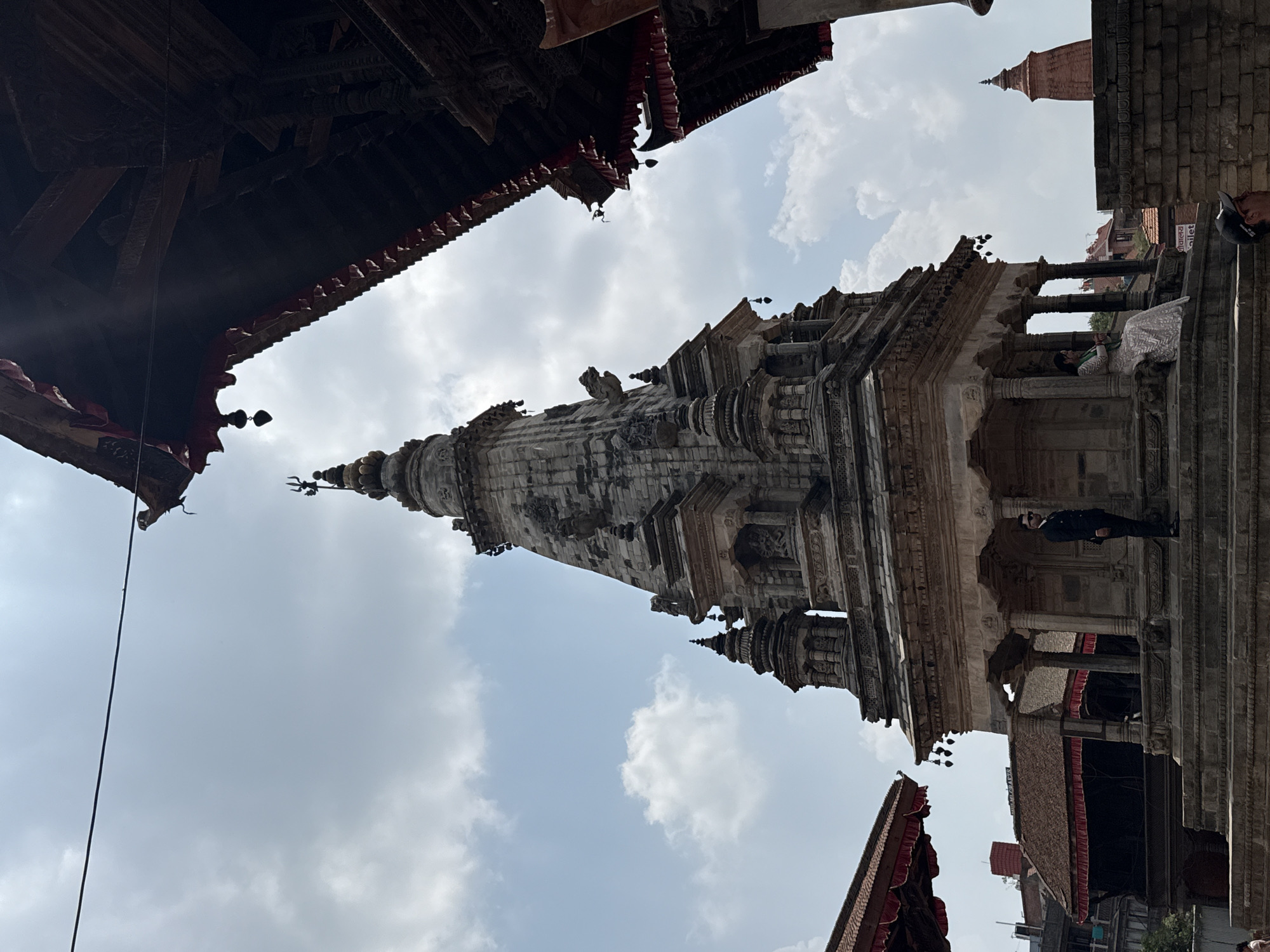A low-angle shot depicts a stone temple tower in Bhaktapur, Nepal, against a backdrop of a cloudy sky. The temple features multiple tiers with ornate carvings and a spire at the top, and is partially framed by the dark eaves of surrounding structures. A man in dark clothing stands near a section of the temple's base, while another, smaller brick temple tower is visible to the right. Red and white trim is visible hanging off the dark eaves, as well as sections of the surrounding city below.