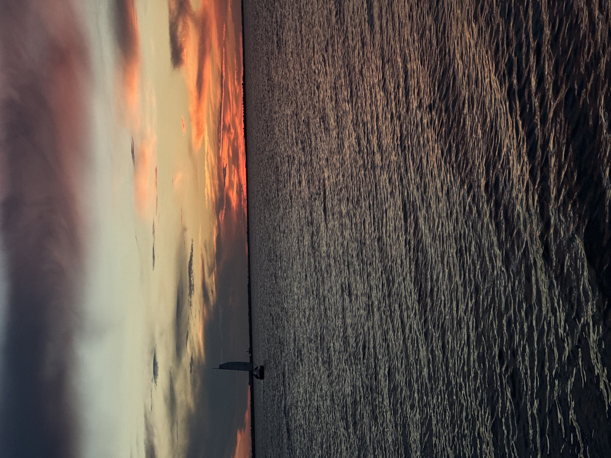 The image depicts a tranquil sunset over a body of water. A small sailboat with two individuals on board is sailing in the distance. The sky is a canvas of vibrant colors, with hues of orange, pink, and purple painting the clouds. The water is calm, with gentle ripples reflecting the warm glow of the sunset. The scene evokes a sense of peace and serenity.