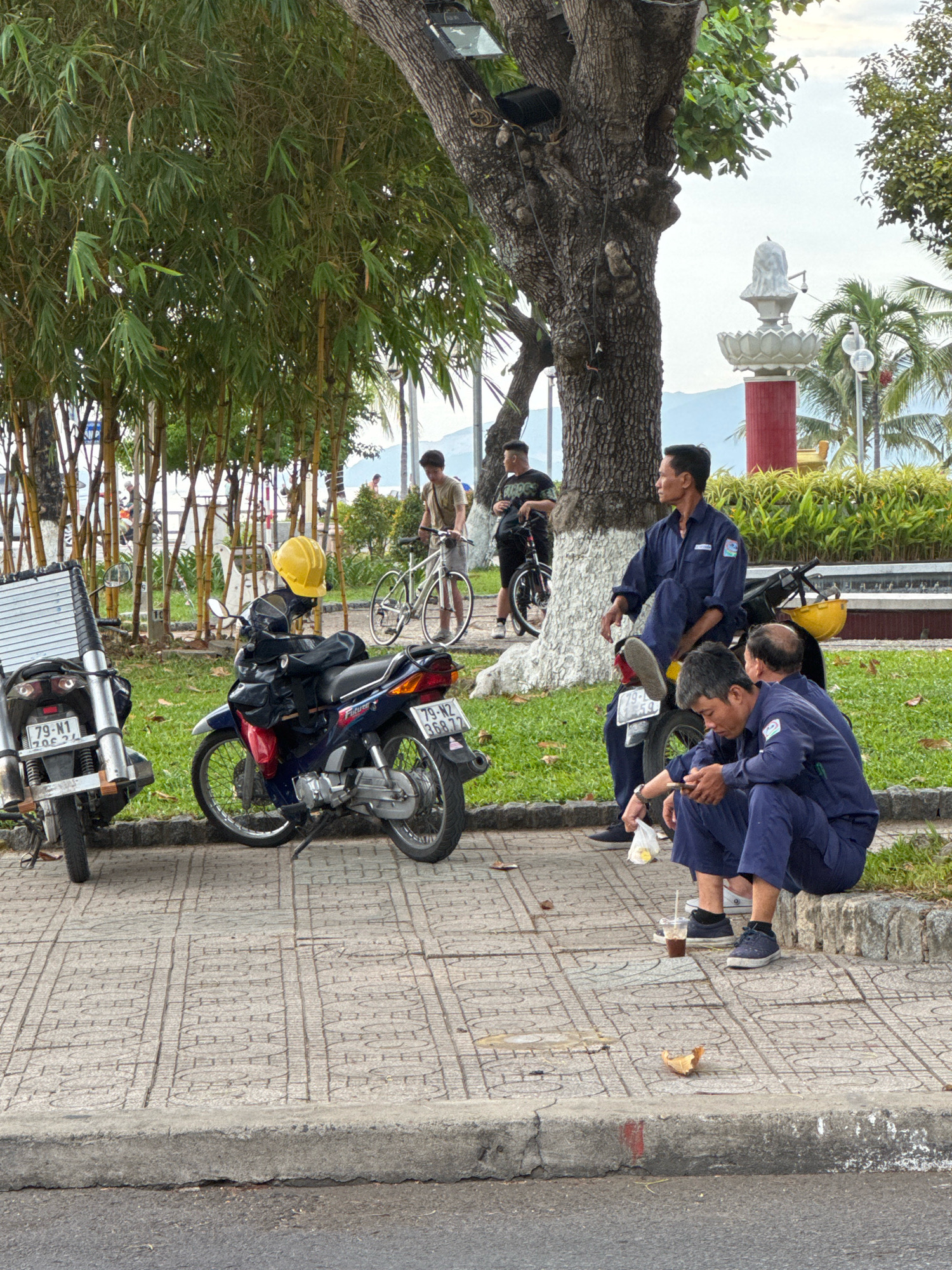 This image captures a scene in Nha Trang, Vietnam, likely along a waterfront promenade or park area. The foreground features a sidewalk with a distinct patterned tile design and a concrete curb separating it from a road. On the sidewalk, three men in blue work uniforms are seated or resting, with two of them appearing to be conversing and holding drinks in disposable cups. One man is looking at his phone. Two motorbikes are parked on the edge of the sidewalk, one black and silver with a storage rack, and a blue and red one with a yellow helmet on its handlebars. In the background, a large tree with rough bark dominates the mid-ground, with a smaller tree to its right. Beyond the trees, there is lush green foliage, including a dense bamboo grove on the left and manicured bushes and palm trees to the right. A white statue on a red pedestal is visible further back. In the distance, a calm body of water and possibly mountains or hills are discernible under a bright sky, suggesting a coastal or bayside setting. A man with a bicycle is also visible in the background, as are other distant figures and structures. The overall impression is one of a public outdoor space where people are taking a break.