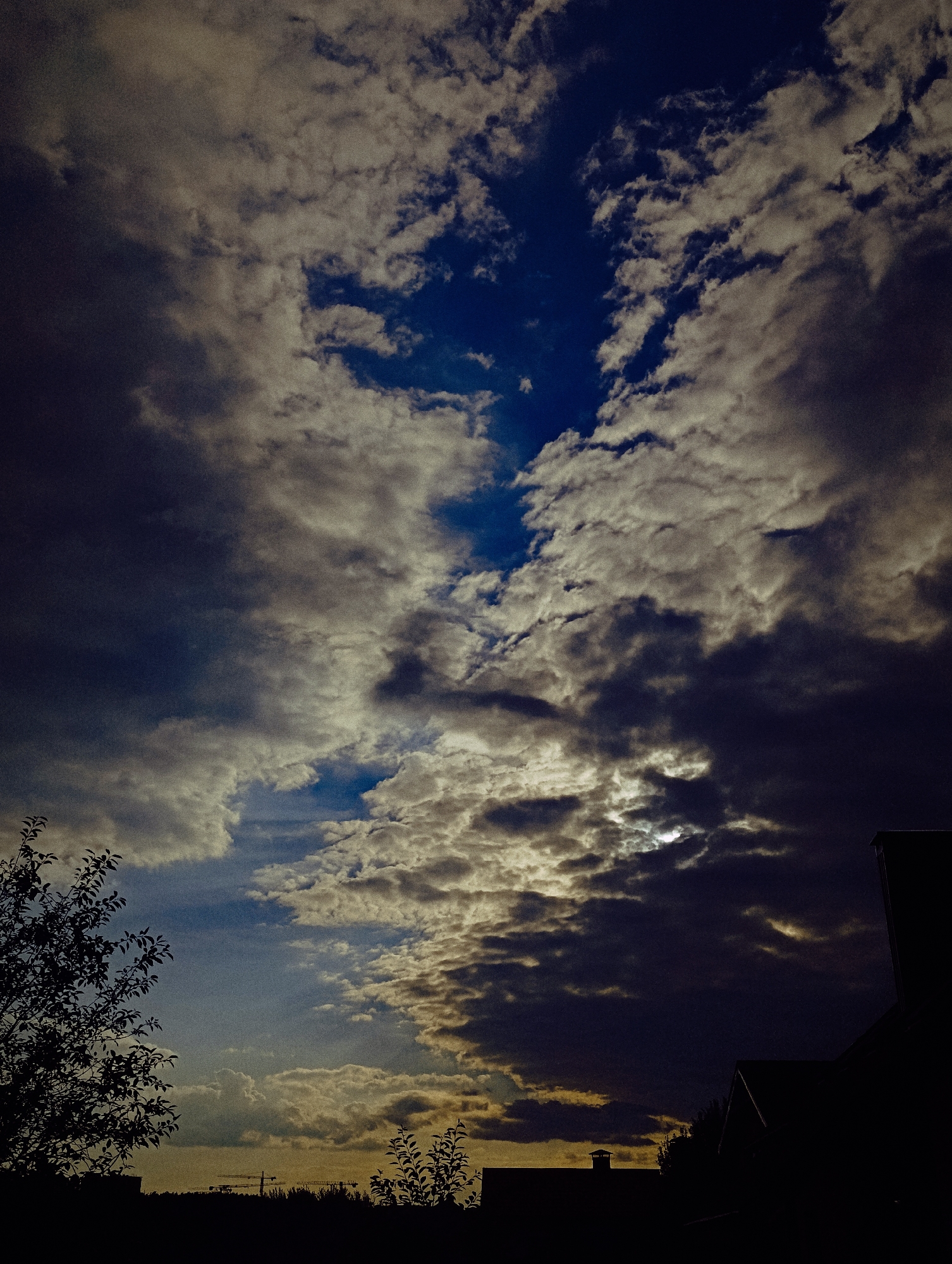 The image depicts a dramatic sky filled with a mix of dark and light clouds. A sliver of the sun is visible peeking through the clouds in the upper right quadrant of the image. The lower portion of the image is silhouetted against the sky, showing the dark shapes of trees and buildings, including a chimney. There are also construction cranes visible in the distance. The overall impression is of a moody, overcast sky with hints of sunlight breaking through. The location is stated as Unknown, Unknown.