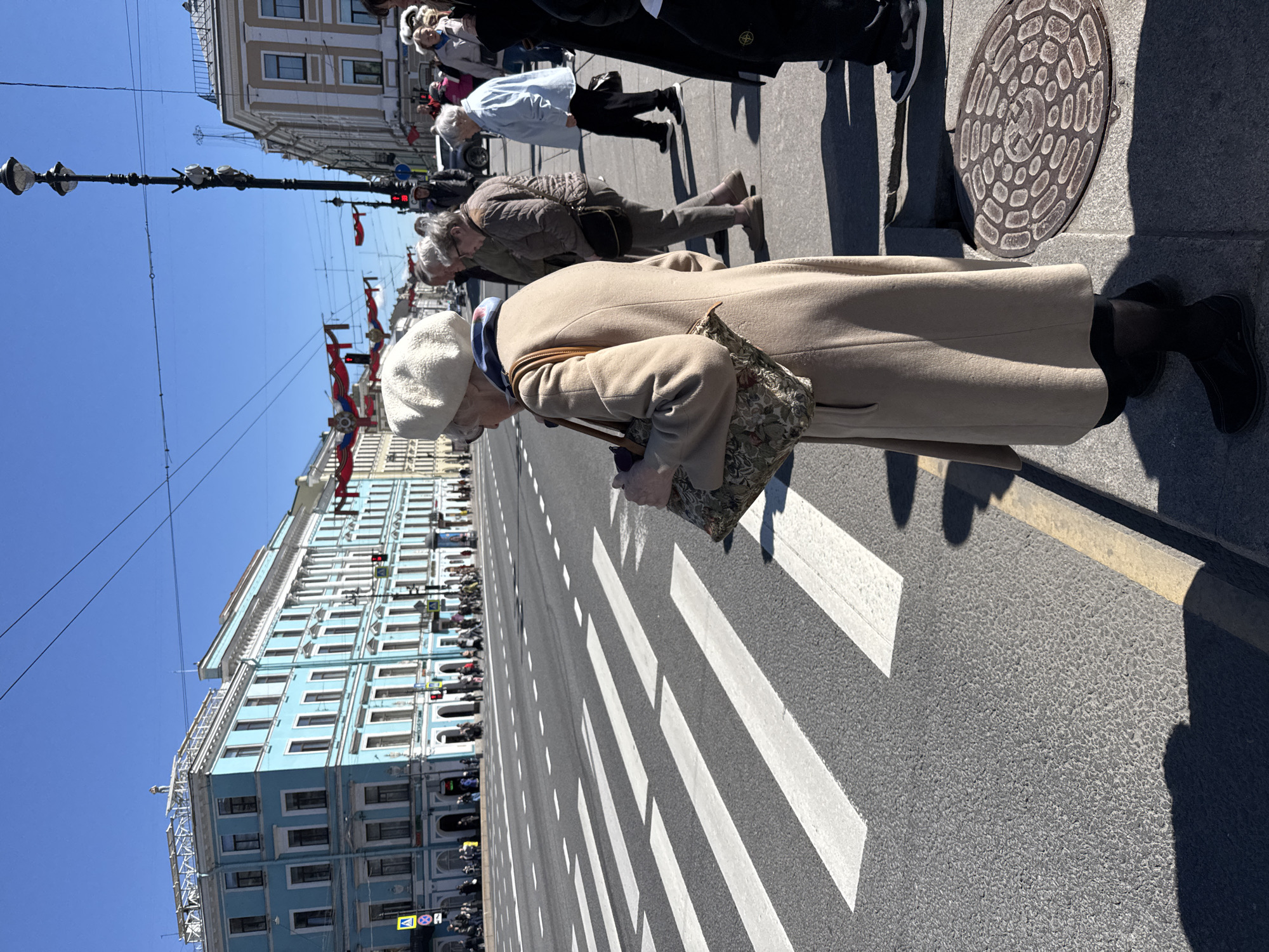 The image captures a street scene in Saint Petersburg, Russia, specifically Nevsky Prospekt. An elderly woman wearing a beige coat and a white beret is crossing the street on a pedestrian crosswalk. Several other people, including older individuals, are also walking in the same direction. The blue building with many windows is visible along the street. Red flags are strung between lampposts. The sky is clear and blue. A manhole cover is located on the sidewalk.
