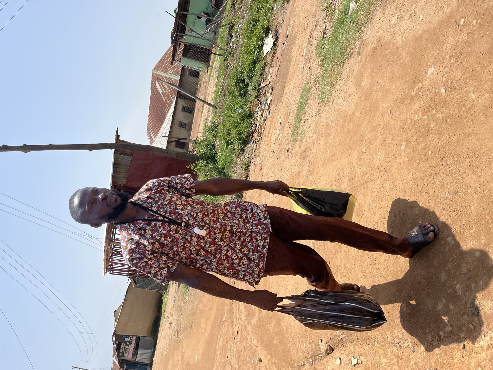 A man is walking on a dirt road with two black bags, one in each hand. He is wearing a floral patterned shirt and brown pants. The man is looking at the camera with a slight smile. The sun is shining, casting a shadow of the man on the road. The background shows some buildings with corrugated metal roofs and some greenery. The scene appears to be in a rural or semi-rural area of Jos, Nigeria. The overall impression is one of a casual daytime activity in a hot climate. There is no visible text in the image.