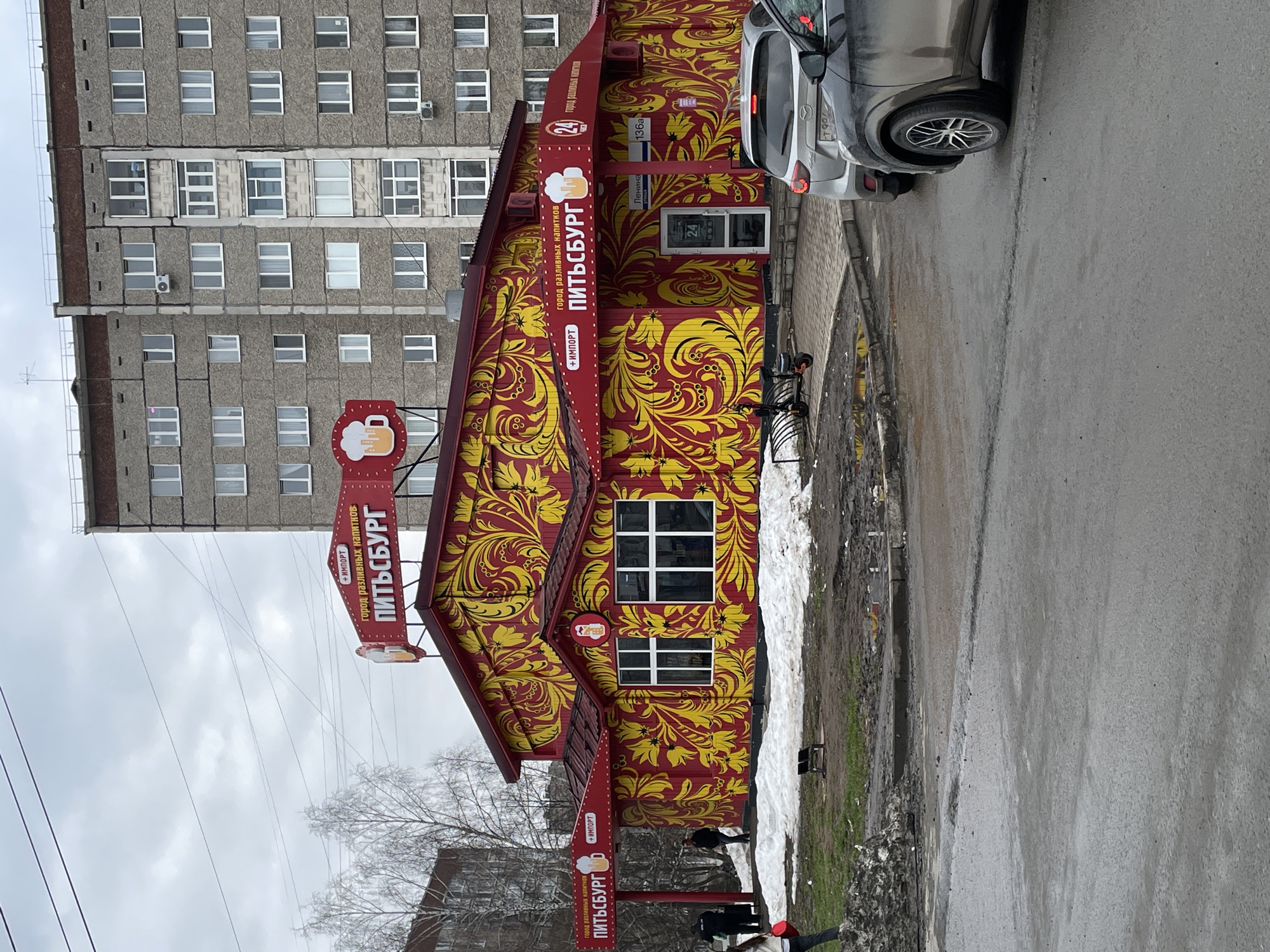 The image shows a red and yellow building in Izhevsk, Russia, with "ПИТЬСБУРГ" (Pittsburgh) written on the side. The name is accompanied by the phrase "город разливных напитков" (city of bottled beverages). A sign indicates that the establishment is located on Lenina Street at number 136a. The shop is open 24 hours. A gray apartment building is in the background. There is a car parked along the curb and some slush on the ground. People are standing outside.