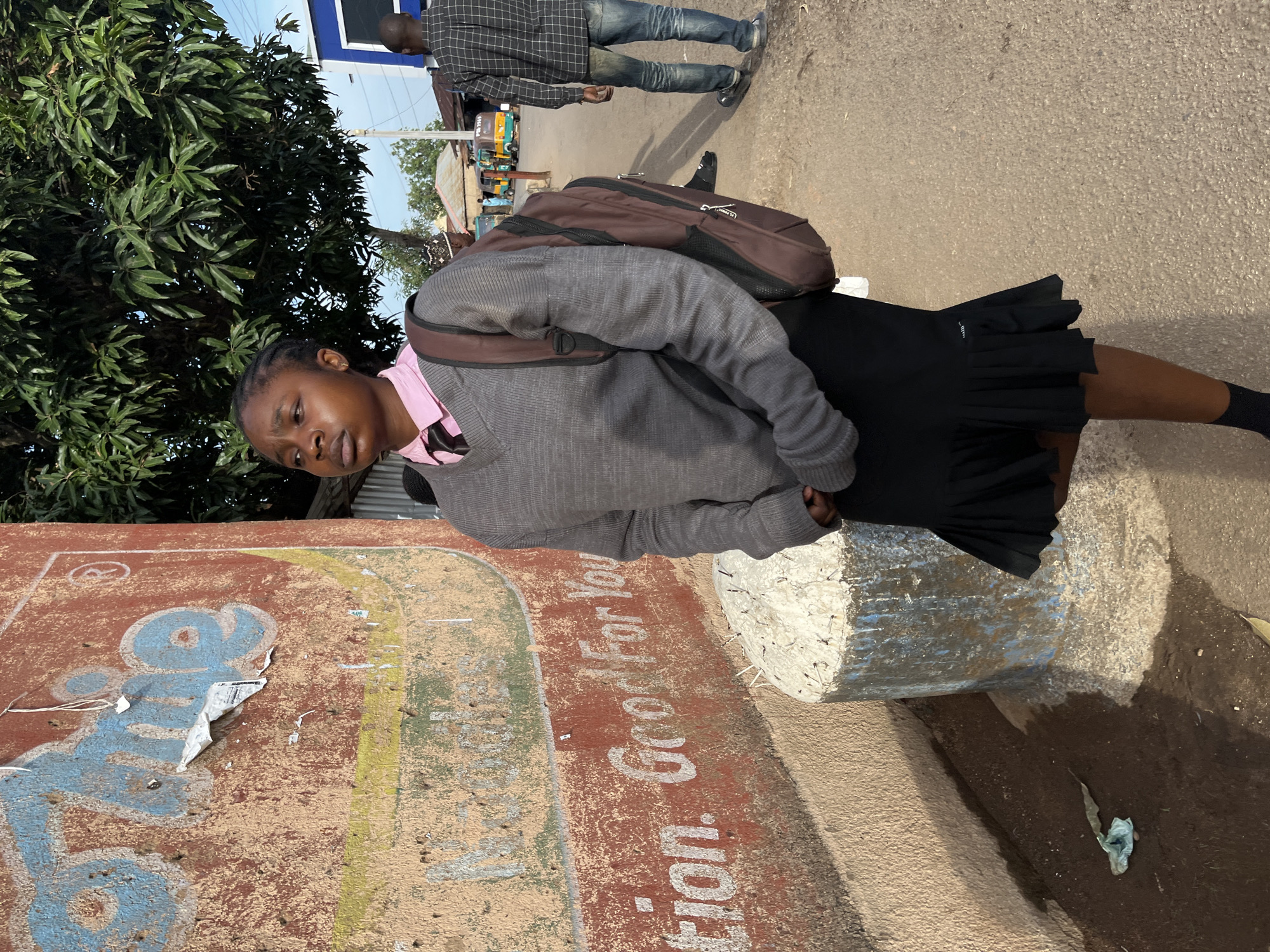 A young woman stands outdoors in what appears to be an urban or semi-urban street setting in Jos, Nigeria, during bright daylight. She has dark skin and braided hair, wearing a pink collared shirt beneath a grey long-sleeved sweater, a black pleated skirt, and dark socks. A brown backpack is slung over her shoulders. She looks slightly downwards with a neutral or pensive expression, her hands seemingly tucked into her skirt or waistband.
In the background, a man in a plaid shirt and blue jeans walks away from the viewer on a paved road. Further down the road, distant vehicles, possibly auto-rickshaws, and other people are visible. To the left of the young woman is a large, leafy green tree, likely a mango tree, its dense foliage filling the upper left of the frame.
The ground she stands on is a mixture of dirt and a paved area. Next to her on the right is a rough concrete barrier or post, painted white and blue, with some rebar poking out from the top. To her left, there's a rustic wall or low structure with a reddish-brown base and faded blue and yellow painted elements. Partially visible text on this wall reads "tion. Good For You". The scene captures a candid moment of everyday life.