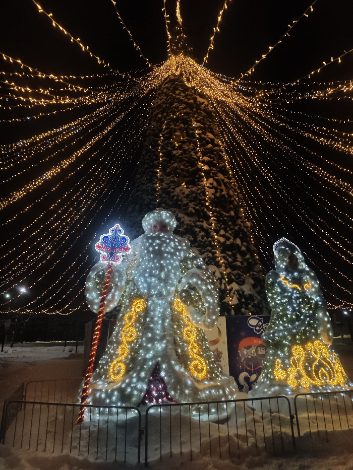 The image displays a vibrant nighttime scene adorned with festive winter holiday decorations. In the foreground, two large, illuminated figures resembling Ded Moroz (Father Frost) and Snegurochka (Snow Maiden) stand on snow-covered ground. They are enclosed within a low, dark metal fence.
The larger figure on the left, representing Ded Moroz, is brightly lit with numerous white string lights, forming his body and a long, white beard. His coat features distinct, ornate yellow and orange scroll-like patterns. He holds a staff with a prominent, intricate blue and red top ornament, which includes heart shapes and some small, illegible text. To his right, the smaller figure, likely Snegurochka, is similarly illuminated with white string lights and adorned with matching yellow and orange swirling patterns on her attire.
Dominating the background is a very tall, dark evergreen tree, possibly covered in a light dusting of snow. This tree is profusely decorated with countless small, warm-toned golden string lights, which cascade downwards along its branches. Extending from the top of this central tree, a spectacular canopy of warm golden string lights stretches outwards across the dark night sky, creating an immersive, star-like effect over the entire area.
The ground surrounding the figures and the tree is covered in a layer of pristine white snow, indicating a winter setting. Behind the illuminated figures, a colorful banner or sign is partially visible. It features a prominent red star or emblem and some Cyrillic text, though most of it is obscured. The characters "Улыб" (Ulyb) are faintly discernible on the sign. The overall impression is one of a magical, festive winter wonderland, likely a public holiday display.