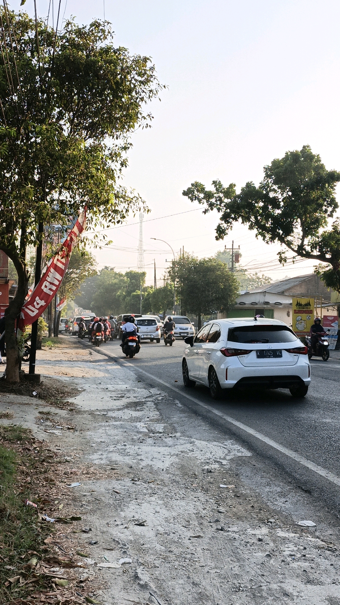 The image depicts a street scene in the city of Sumbring, Indonesia, under a bright, hazy sky. The road is moderately busy with traffic, primarily consisting of motorcycles and cars. In the foreground, a white hatchback car is prominently featured, facing away from the camera and traveling on the right side of the road. A red and white banner, likely a flag or advertisement, hangs from a pole on the left side of the image, partially obscuring a portion of the street and sidewalk.
The sidewalk on the left is unpaved and appears somewhat weathered, with patches of dirt, scattered leaves, and debris. Lush green trees line the sidewalk, casting shadows and adding to the natural elements of the scene. On the right side of the road, buildings with simple architecture and some visible signage are present, suggesting a commercial or residential area. Power lines and a distant telecommunications tower are visible in the background, indicating the presence of infrastructure.
Several individuals are on motorcycles, traveling in the same direction as the white car. One motorcyclist in the foreground is wearing a white helmet and a blue shirt, while others in the midground are seen in various attire. The overall impression is one of everyday urban life in a tropical or subtropical environment, with a mix of natural and man-made elements. The lighting suggests it might be morning or late afternoon, given the soft, diffused sunlight.