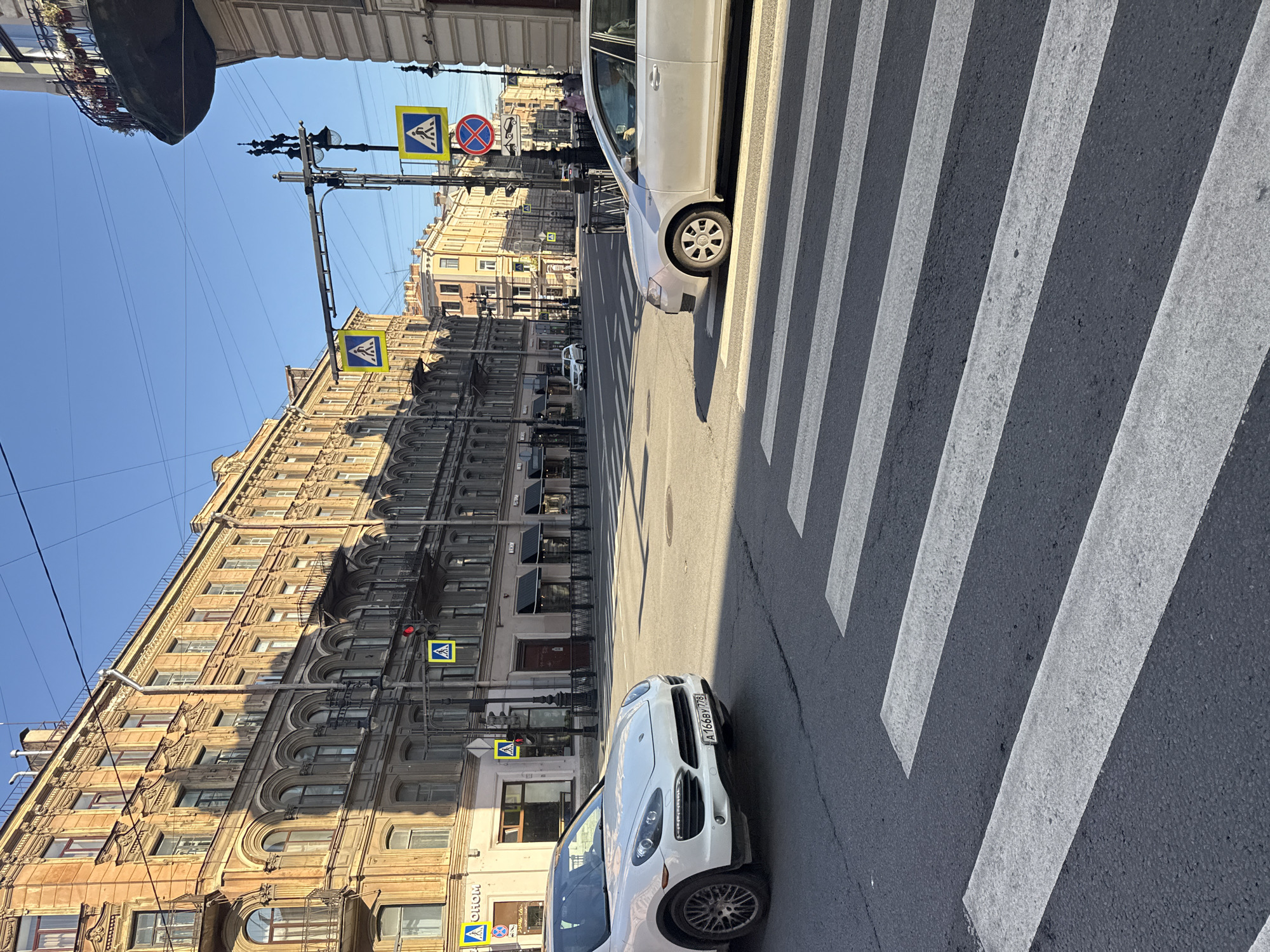 The image depicts a city intersection in Saint Petersburg, Russia, characterized by ornate 19th-century architecture and clear, sunny weather. A white Porsche Cayenne SUV is positioned in the foreground, facing left, and a silver sedan is in the mid-ground, facing right. Both vehicles are on the road, with pedestrian crossings visible in the foreground and mid-ground. Several traffic signals and pedestrian crossing signs are present, indicating a regulated intersection. The buildings lining the street feature arched windows, balconies, and detailed facades, typical of historic St. Petersburg. The sky is clear and blue, with some electrical wires crisscrossing overhead. The overall scene captures a moment of urban life in a historically rich European city.