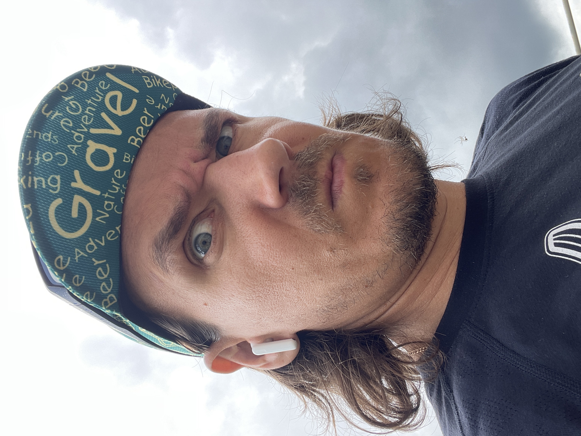 The image is a rotated, close-up selfie of a man, captured from a low-angle perspective with the sky overhead. The man's head is tilted to his left, and he looks directly into the camera with a neutral expression.
The subject is a man with long, wavy brown hair extending past his ears, a short brown beard, and a mustache. He wears a teal green cycling cap with various words printed in yellow and white, including prominent "Gravel," and smaller words like "Adventure," "Nature," "Beer," "Friends," "Coffee," "Hiking," and "Bike." He is also wearing a dark blue or black t-shirt. A white wireless earbud is visible in his right ear.
The background consists primarily of a heavily overcast, grey sky with visible cloud formations, indicating an outdoor setting. In the upper right quadrant of the image, against the cloudy sky, a small, light-colored insect, possibly a spider or moth, is visible.
The image appears to be a personal photograph or selfie, possibly taken during an outdoor activity such as cycling, as suggested by the cap's design. The weather is cloudy daytime.