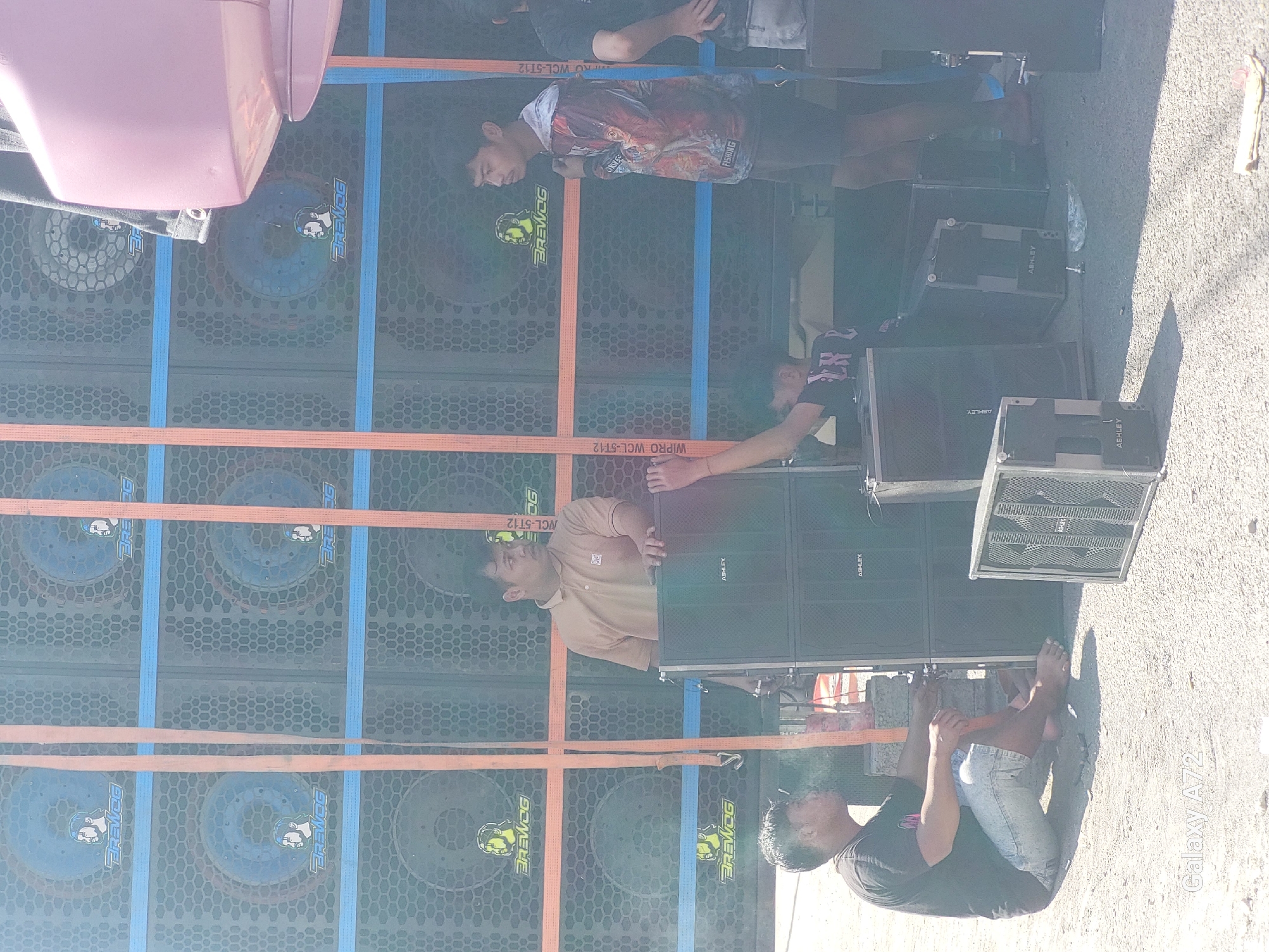 In Sengguruh, Indonesia, a group of individuals are gathered around a large speaker setup. The speakers are stacked vertically and horizontally, with branding visible on some of them. The scene appears to be outdoors, with a rough, gravelly surface underfoot.
Three men are clearly visible in the foreground. One man, wearing a dark t-shirt and light-colored shorts, is sitting on the ground, facing away from the camera. He appears to be interacting with the speaker equipment. Another man, in a light brown polo shirt, is standing and looking towards the speakers. A third man, wearing a patterned long-sleeved shirt, is standing to the right, looking in the same direction as the man in the polo shirt. The legs of another individual are visible to the far right, suggesting there may be more people present or partially obscured.
The speaker units are prominent objects in the scene, with various sizes and configurations. Some of the speakers have a hexagonal mesh grille. Straps, possibly for securing the equipment, are visible running vertically and horizontally across the speaker stacks. The overall impression is one of an event setup or a sound system being prepared or managed. The lighting suggests it is daytime.