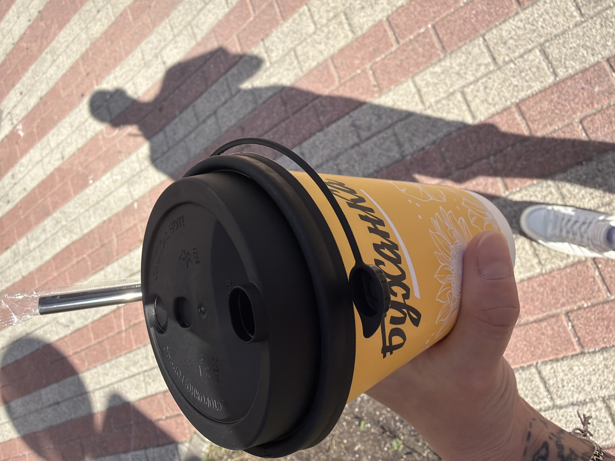 The image shows a person's hand holding a yellow disposable coffee cup with a black lid. The cup has the brand name "Буханка" written in white cursive script, along with a sunflower illustration. A metal straw is visible protruding from the lid, enclosed in plastic packaging. The background consists of a paved walkway with red and beige rectangular tiles arranged in a herringbone pattern. A shadow of a person, presumably the one holding the cup, is cast on the walkway. The scene appears to be outdoors, possibly in a public area of Moscow, Russia, given the Cyrillic script on the cup.