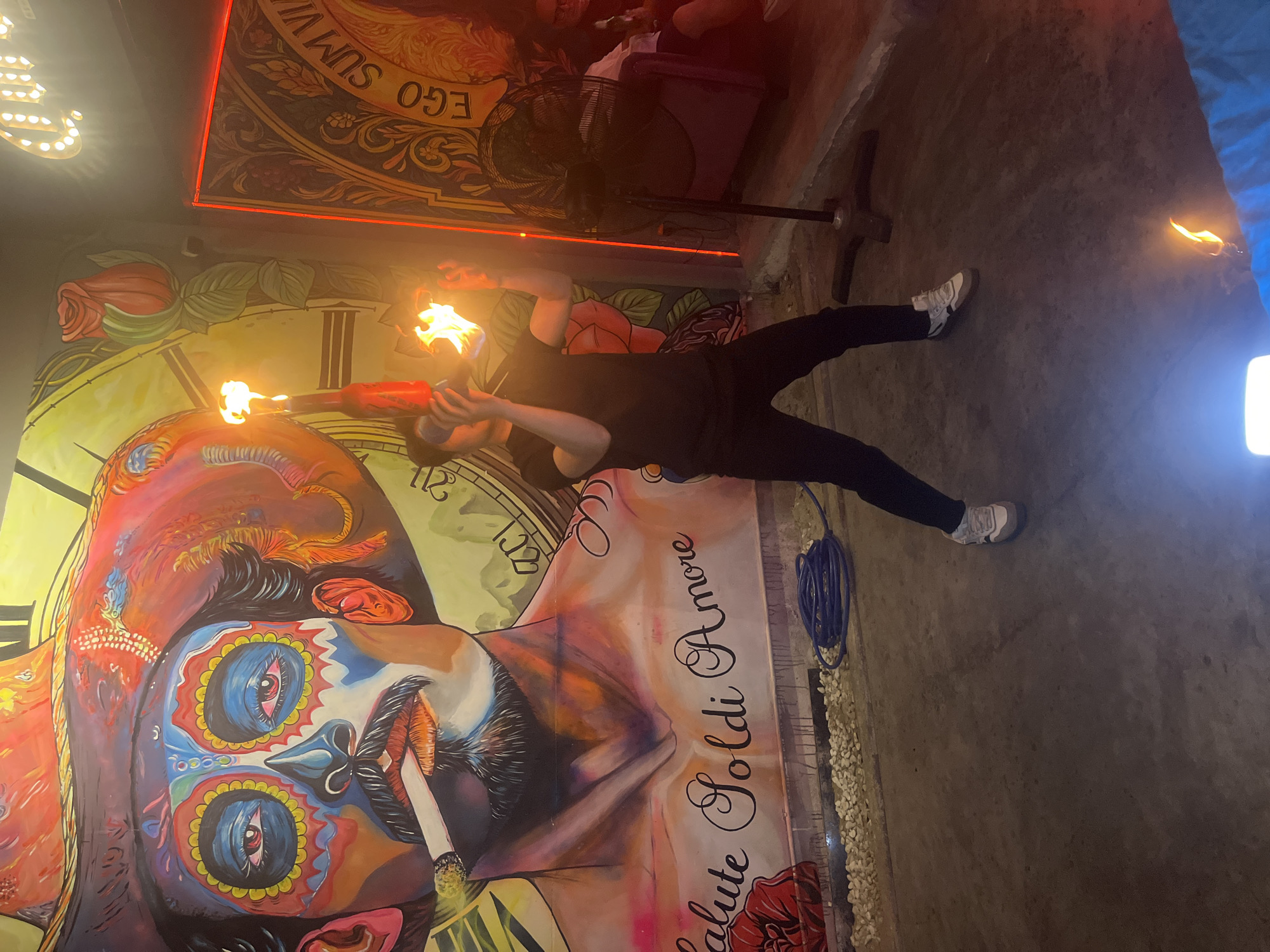 In Đà Nẵng, Vietnam, a performer in a black t-shirt and pants is seen juggling two flaming objects in front of a large mural. The mural depicts a man's face with a sombrero, adorned with colorful patterns resembling face paint, and a cigarette in his mouth. Below the face, the words "salute Soldi Amore" are written. The performer's actions are illuminated by the flames of the objects they are juggling. To the right of the performer, a large black industrial fan stands on a tripod. Behind the performer and to the right, a portion of a wall is visible with a framed artwork and a neon red light strip. The floor appears to be concrete, with a coiled blue hose lying on the ground in front of the mural. In the background, other individuals are present, one of whom is sitting on a couch. The overall setting suggests an indoor or outdoor entertainment venue.