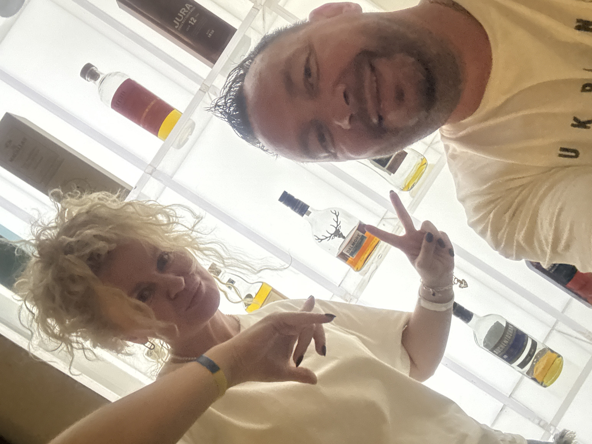 This is a selfie taken by a man and a woman in a brightly lit space, possibly a store or a bar, featuring shelves filled with bottles. The man, with dark, spiky hair and a wide smile, is in the foreground, wearing a white t-shirt with "UKR" visible on it. The woman, with curly blonde hair, is to his left, wearing a light-colored t-shirt and a yellow and blue bracelet on her wrist. She is holding up two fingers in a peace sign. In the background, various bottles are displayed on white shelves, including what appears to be whisky bottles, some with labels such as "JURA 12". The lighting creates a high-key effect, making the background appear very bright. The overall impression is of a casual, happy moment captured in what seems to be a retail or hospitality setting. No specific details about the city of Nanhai District, China are discernable from the image itself.