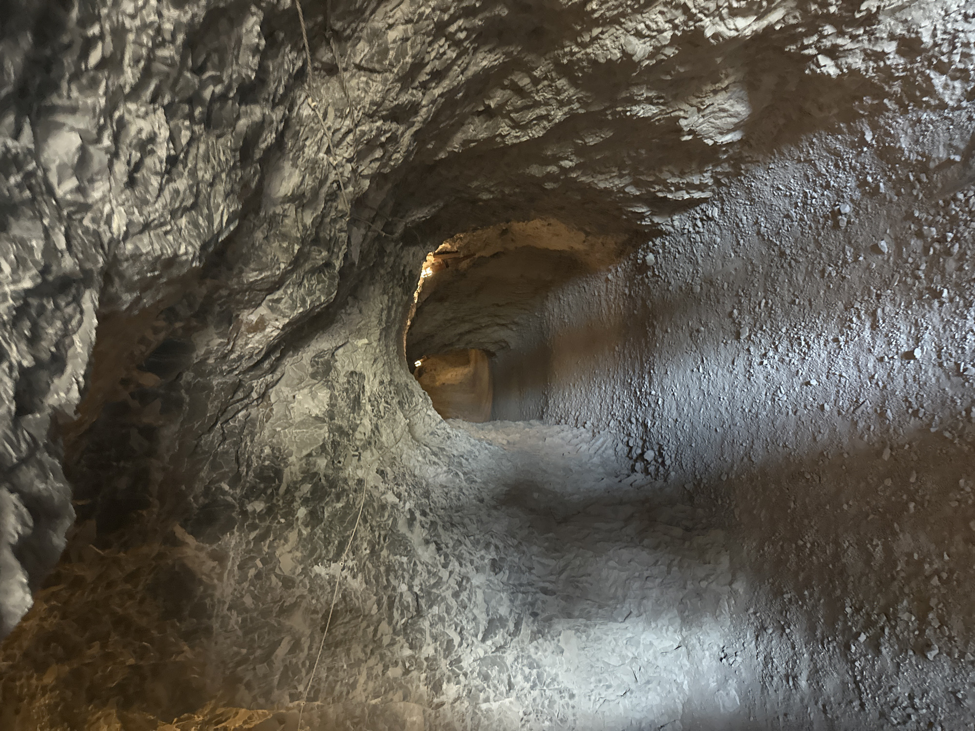 The image displays an illuminated subterranean passage or tunnel located in Novo-Zubutli, Russia. The passage is defined by rough, unhewn rock surfaces forming the walls and ceiling, exhibiting a textured appearance of varying shades of gray, off-white, and earthy brown. The floor consists of uneven, compacted gravel and small stones, indicating a walking path. Artificial light sources, situated deeper within the tunnel, emit warm yellow-orange illumination, casting shadows and highlighting the rugged contours of the rock, particularly along the right-hand wall and further into the receding distance. A thin, light-colored rope or wire is visible spanning horizontally along the upper left section of the tunnel wall. The passage appears to narrow and curve gently to the right as it extends into the background. There are no individuals, specific objects beyond the rope/wire, or discernible actions present in the scene.
