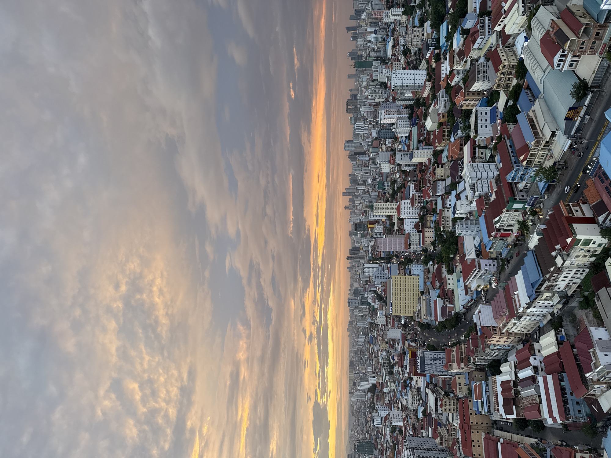 The image is an aerial, wide-angle shot of the city of Khan Mean Chey, Cambodia, taken at dusk. The sky is filled with a dramatic sunset, featuring a gradient of colors from warm oranges and yellows near the horizon to soft grays and blues overhead. Clouds are scattered across the sky, catching the light of the setting sun. Below the sky, a dense urban landscape stretches out, characterized by a mix of low-rise residential buildings with red and blue tiled roofs, mid-rise commercial buildings, and a scattering of taller modern skyscrapers in the distance. Streets are visible, with some vehicle and pedestrian traffic indicated by faint lights and smudges. The overall impression is of a bustling city viewed from a high vantage point during a vibrant sunset.