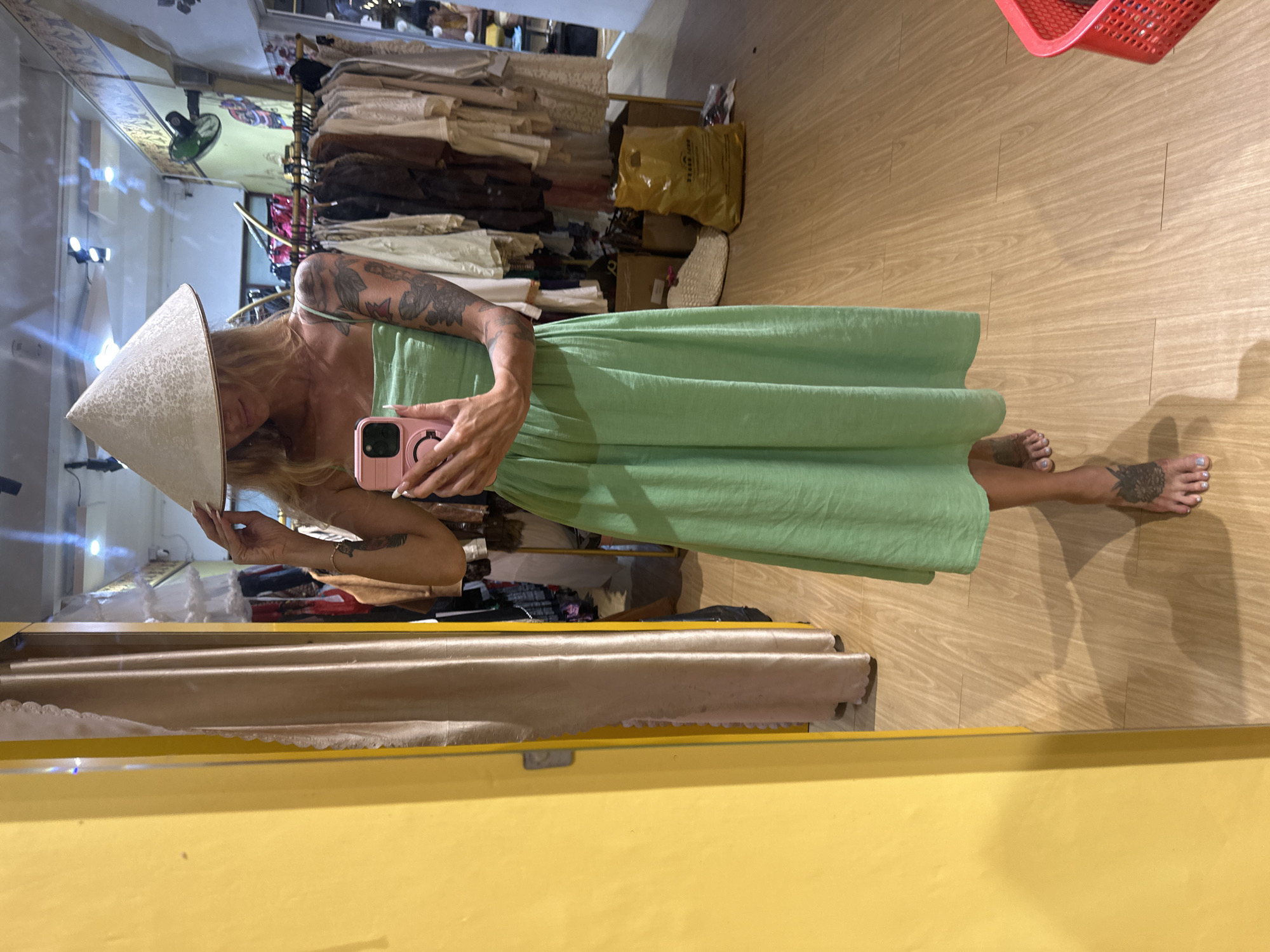 A person stands in a boutique, reflected in a large mirror that dominates the left side of the frame. The person, wearing a conical hat and a green dress, holds up a pink smartphone to take a selfie. The background displays racks of clothing and other shop items. The floor is made of wooden planks, and a yellow wall is visible on the left.