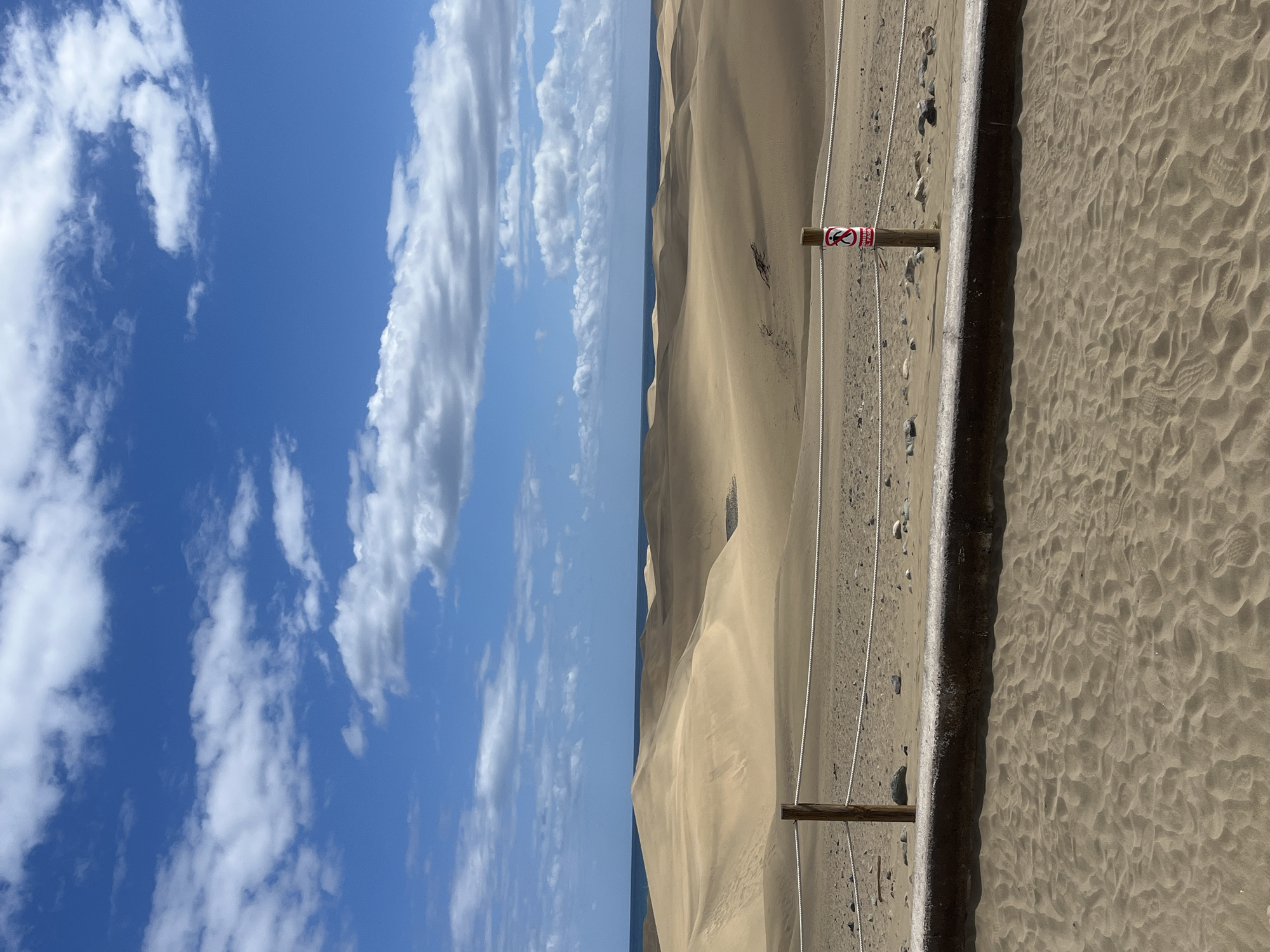 The image shows a bright blue sky with scattered white clouds on the left side, separated from sand dunes on the right side by a thin horizon line. The dunes are tan and sandy, with subtle variations in shading that indicate their undulating form. A barrier made of wooden posts and white ropes runs along the edge of the dunes, with a red and white sign attached to one of the posts. The ground immediately to the right of the barrier is sand, showing many small footprints and textures. This is a view of the Maspalomas dunes in San Bartolomé de Tirajana, Spain.