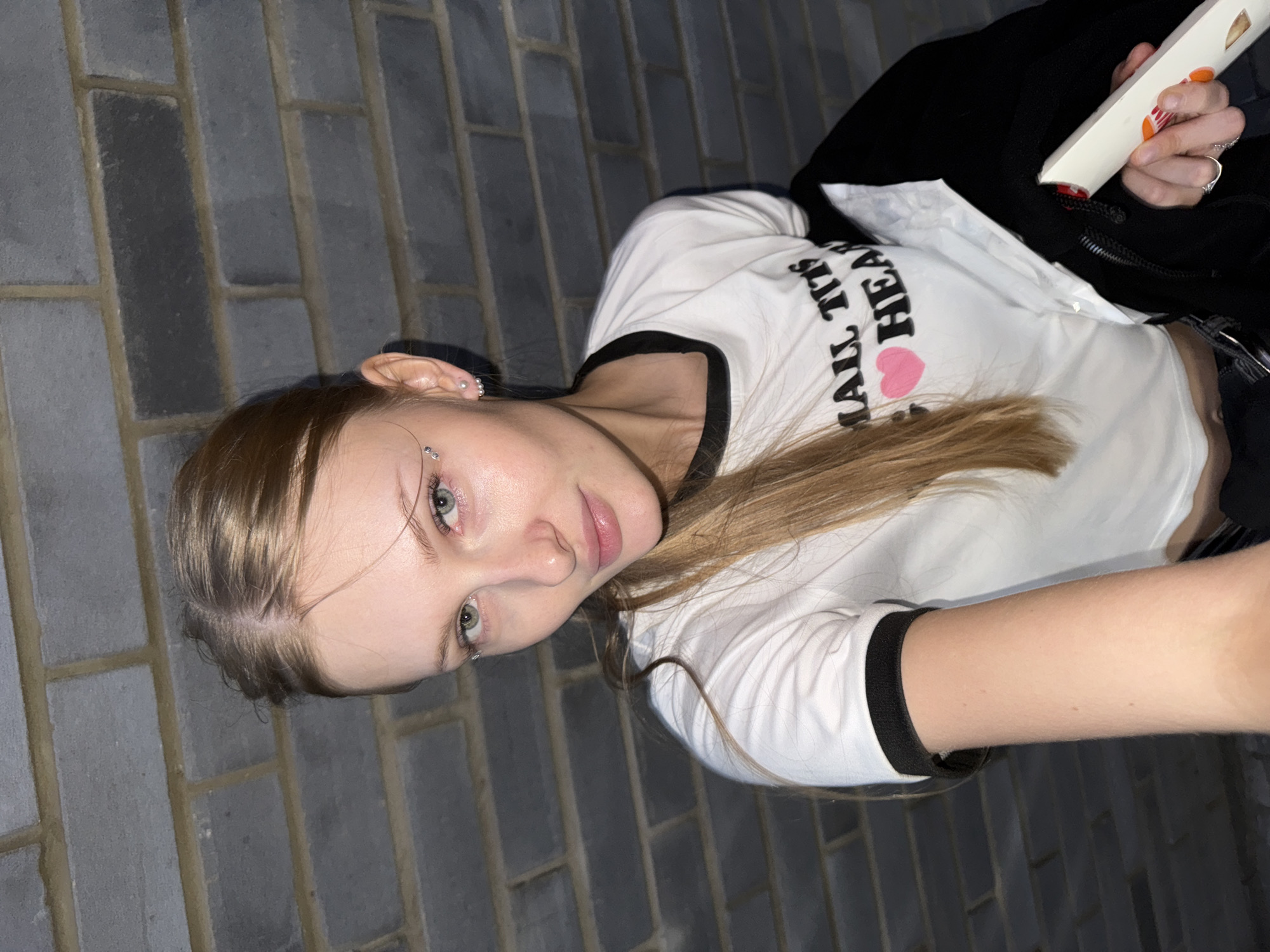 A young Caucasian woman with blonde hair is positioned in the foreground, looking directly at the camera. She is wearing a white cropped t-shirt with black trim that has the text "SMALL TITS HEART" and a pink heart printed on it. She also has a black jacket draped over her left arm and is holding a white object with orange and red accents in her right hand. The background is a brick wall, which appears to be a common architectural element found in the city of Perm, Russia. The lighting suggests it might be evening or an indoor setting with artificial lights. There are no other individuals or significant objects in the frame.