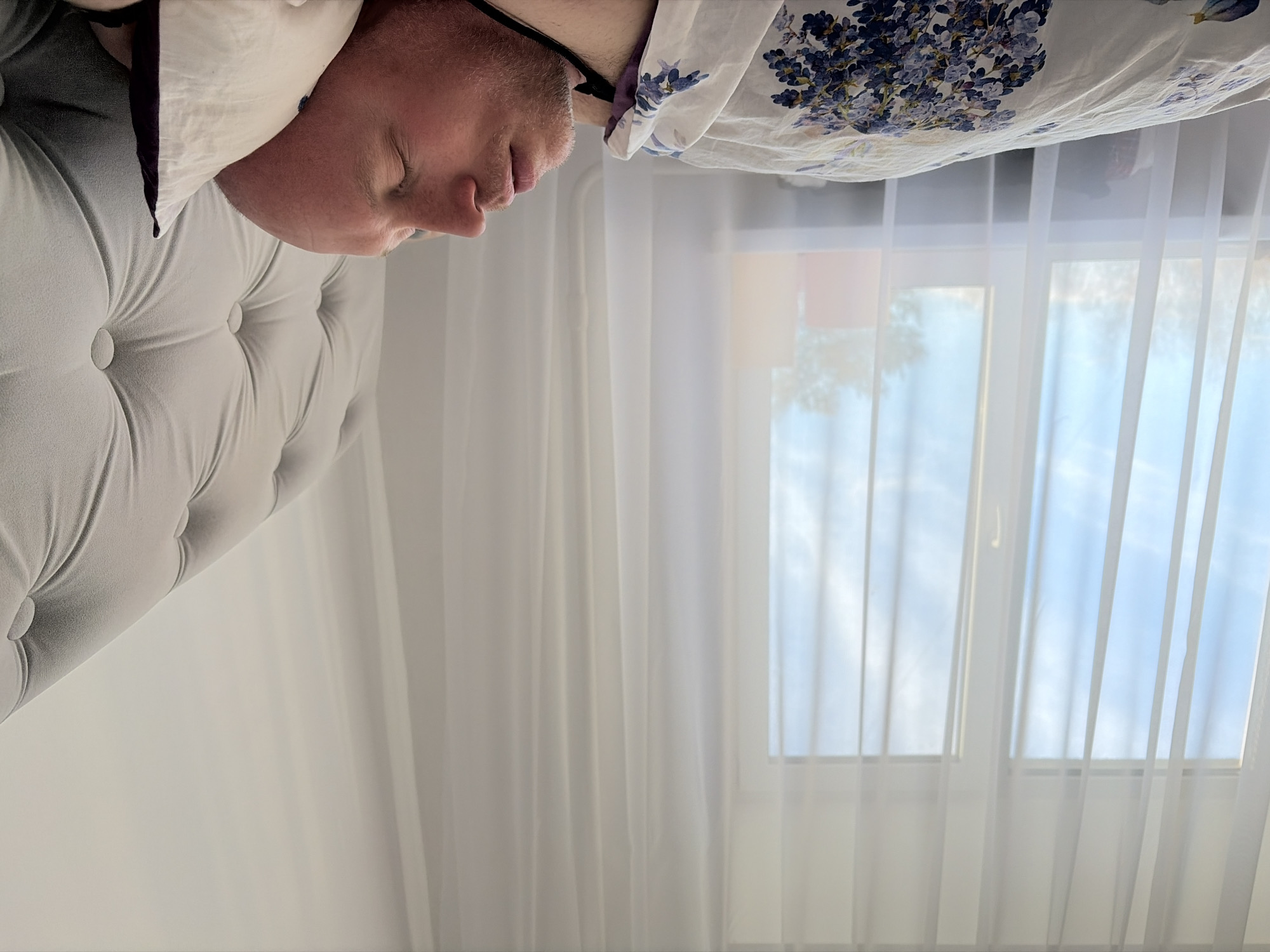 A male individual is depicted sleeping in a bed located in a room within Chaikovsky, Russia. The man, with a bald head and light facial stubble, lies on his back or side, eyes closed, resting on a white pillow with dark trim. He is partially covered by a white duvet featuring a blue and purple floral pattern. A light grey, tufted upholstered headboard is visible to the right. In the background, a large window is partially obscured by sheer white curtains, revealing a blue sky with faint clouds and bare tree branches outside. Several indistinct colored objects, including red and orange, are visible on a surface near the bottom of the window. Natural light illuminates the scene.
