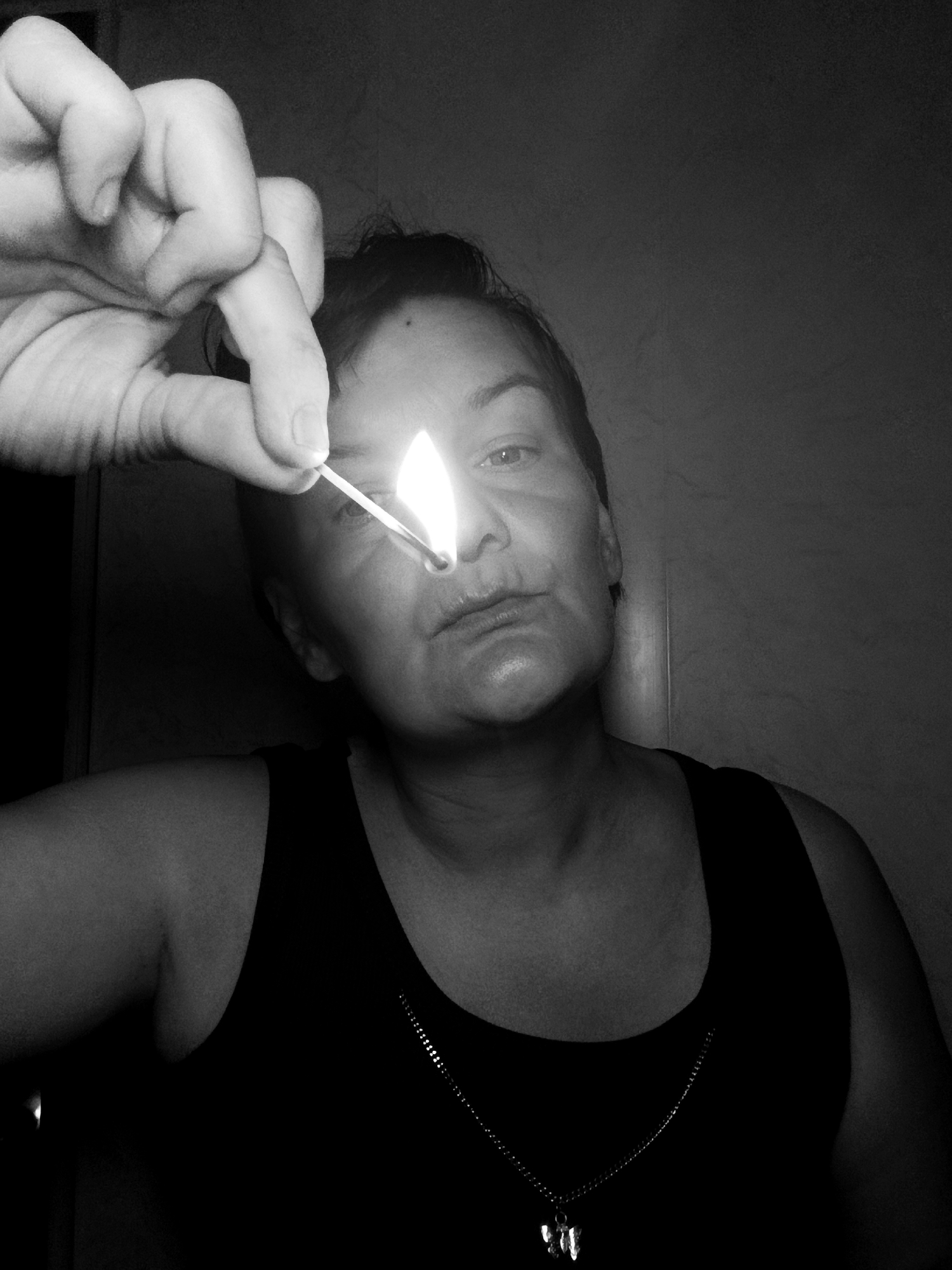 A medium, close-up black and white photograph shows a woman holding a lit match in front of her face. The woman is wearing a dark tank top and a chain necklace. Her face is illuminated by the match. The background is a dark wall. The photo was not identified to be taken in the city of Фоки, Russia.