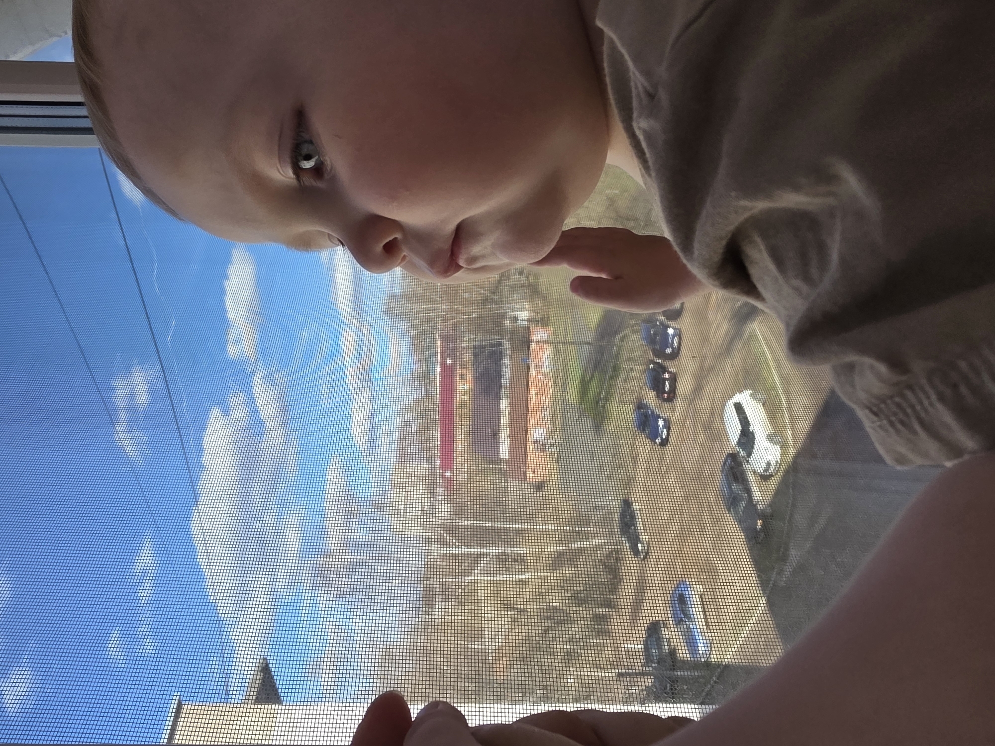 A young child's face peers through a screened window, with their right hand touching their lips. The view through the screen shows a partially obscured outdoor scene, including a blue sky with clouds, buildings, and parked cars. The location appears to be Chaikovsky, Russia.