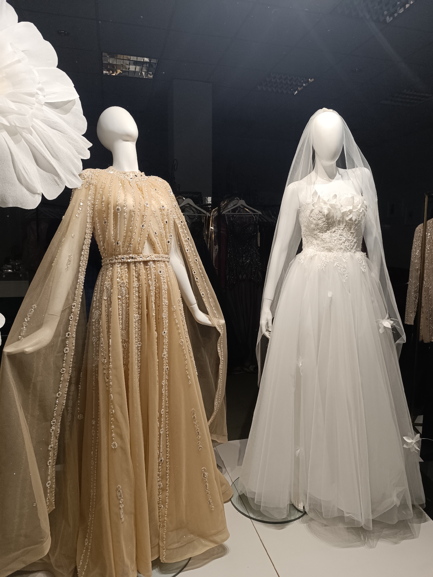 A vertical, eye-level shot shows two mannequins in a storefront display, presumably in an unknown city. The mannequin on the left wears a beaded, gold-colored gown with long, sheer sleeves that extend to the floor. It has a belt around the waist, also beaded. The mannequin on the right wears a white wedding dress with a veil. The dress has floral detailing on the bodice and skirt. A large white flower is to the left of the gold dress. The background is dark and indistinct, but other dresses on hangers can be made out. The floor is a glossy white surface. The mannequins are positioned close together. The scene appears to be taken indoors, likely a boutique or bridal shop.