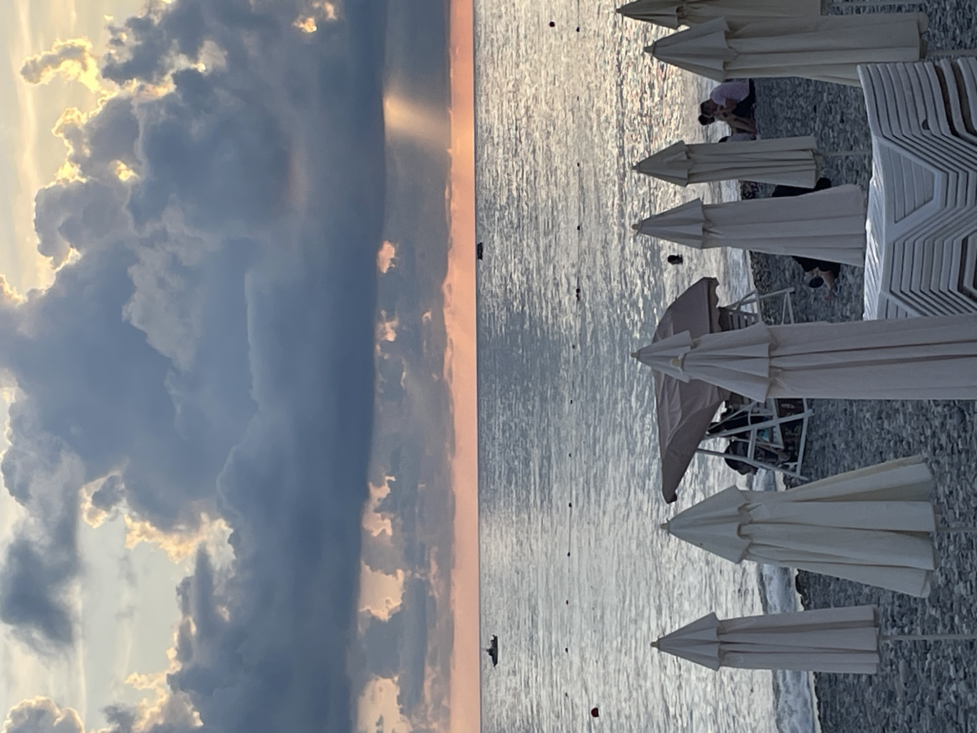 The image captures a serene beach scene at dusk in an unknown city. The sky is filled with dramatic, dark clouds, partially illuminated by the setting sun, which casts rays of light onto the calm sea. Several white beach umbrellas are closed and lined up on the pebbled shore in the foreground. A few people are visible in the distance, some sitting on the beach, others swimming in the ocean. A small boat is also present on the water. The overall atmosphere is peaceful and tranquil, with the natural beauty of the seascape taking center stage.