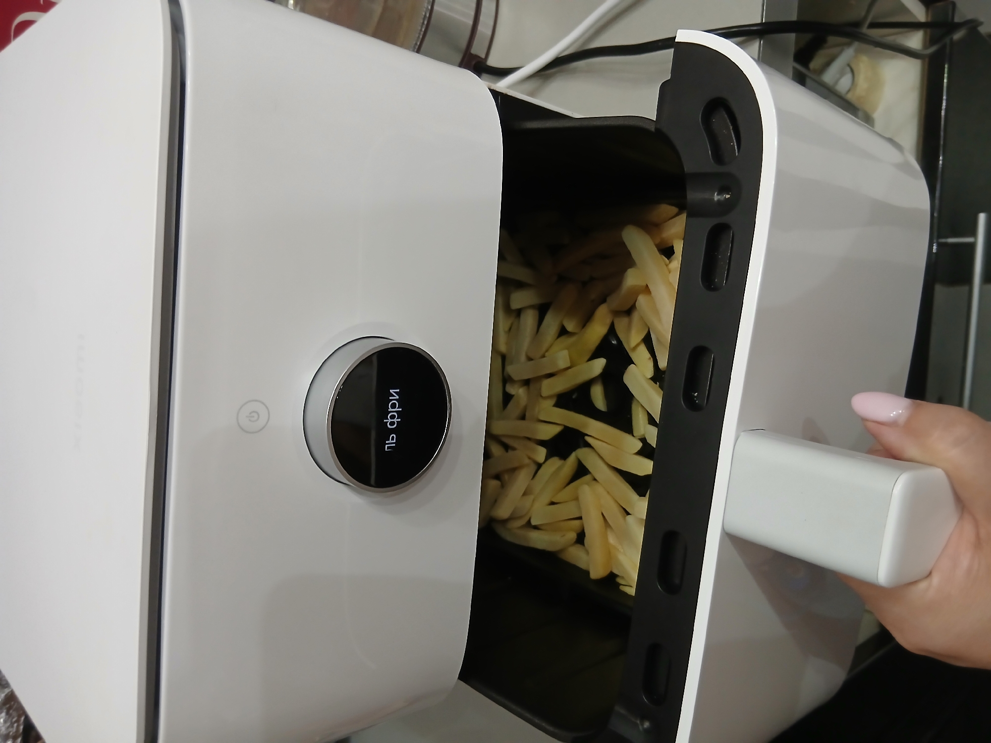 A person's hand, with light pink nail polish, is holding the white handle of an open air fryer basket. The basket is filled with french fries. The air fryer is white with a black interior and a circular control panel displaying the Cyrillic text "пь фри". The scene is indoors.