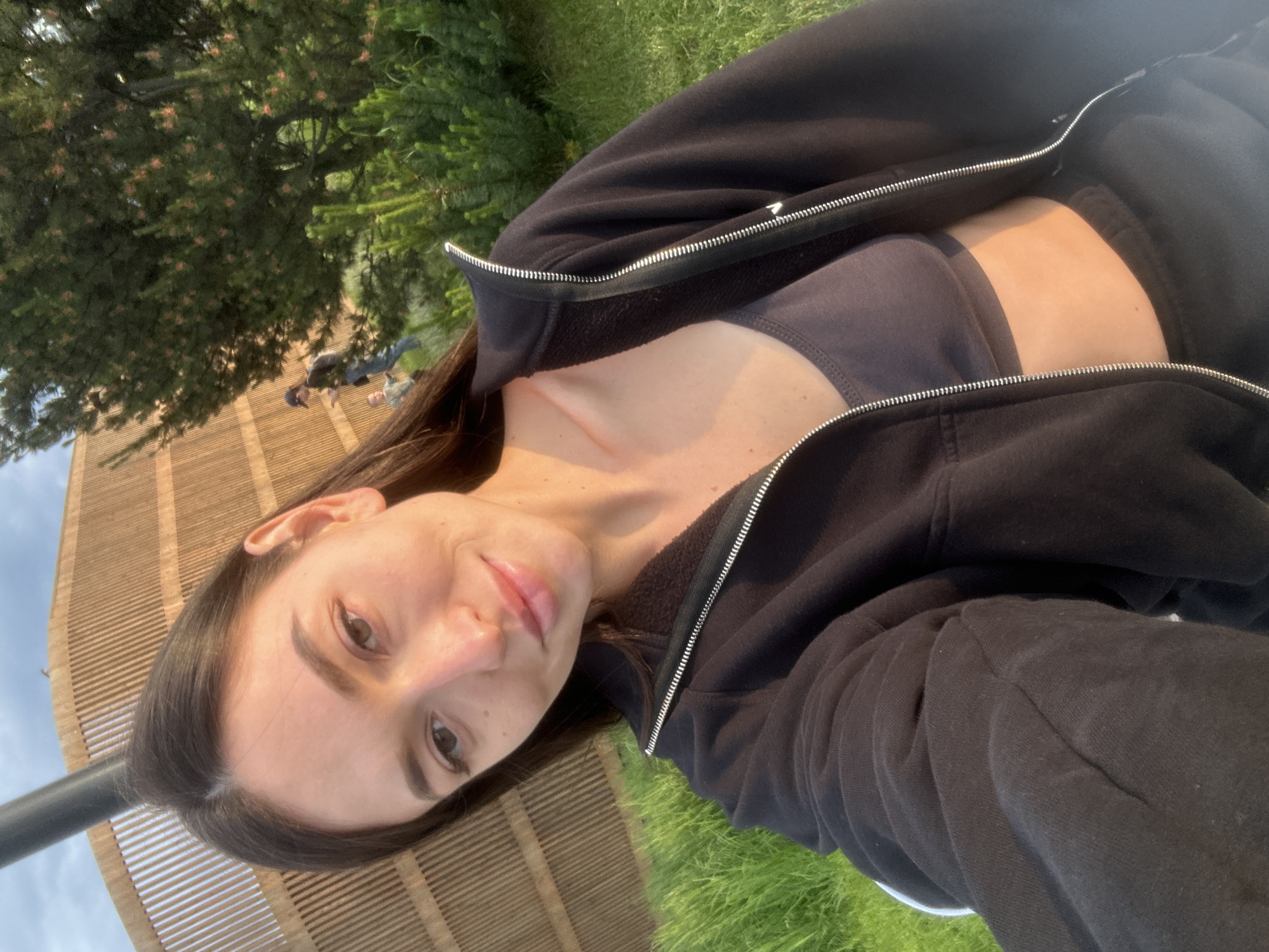 A woman with dark hair takes a selfie outdoors, in what appears to be a park or recreational area. She is wearing a dark zip-up hoodie over a dark sports bra and black pants. The background features a wooden structure with vertical slats, lush green grass, and trees. In the distance, two other individuals can be seen, one appearing to be a child. The lighting suggests it is late afternoon or early evening. There is no specific indication of the location being in Kazan, Russia.