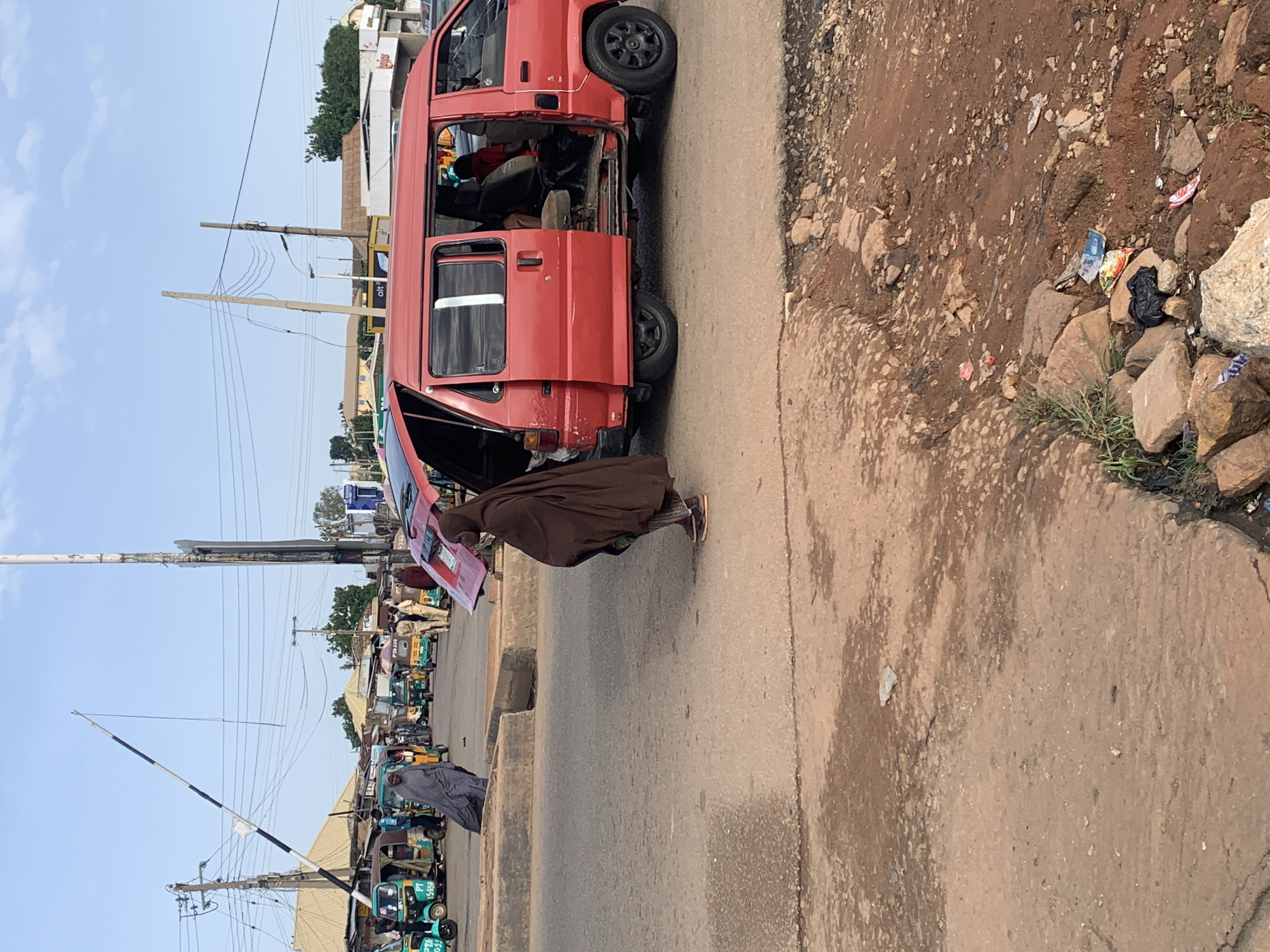 The image depicts a street scene in Gura Top, Nigeria, with a red minivan prominently featured. The minivan has its sliding doors and rear hatch open, revealing its interior and passengers. Two individuals are visible near the minivan: one is a person wearing a brown abaya, standing to the right of the minivan and appearing to be walking away from it. Another person, dressed in a grey abaya, is visible in the background, standing to the left of the minivan.
The street itself appears to be paved with asphalt, showing signs of wear and tear, with a dirt and rocky embankment on the right side of the frame. In the background, several auto-rickshaws (also known as "keke napep") are parked or in motion, indicating a busy transportation hub. Buildings with corrugated metal roofs are visible in the distance, along with utility poles and overhead electrical wires. The sky is clear and blue, suggesting a daytime setting. The scene appears to be a typical urban environment with active commerce and transportation.
