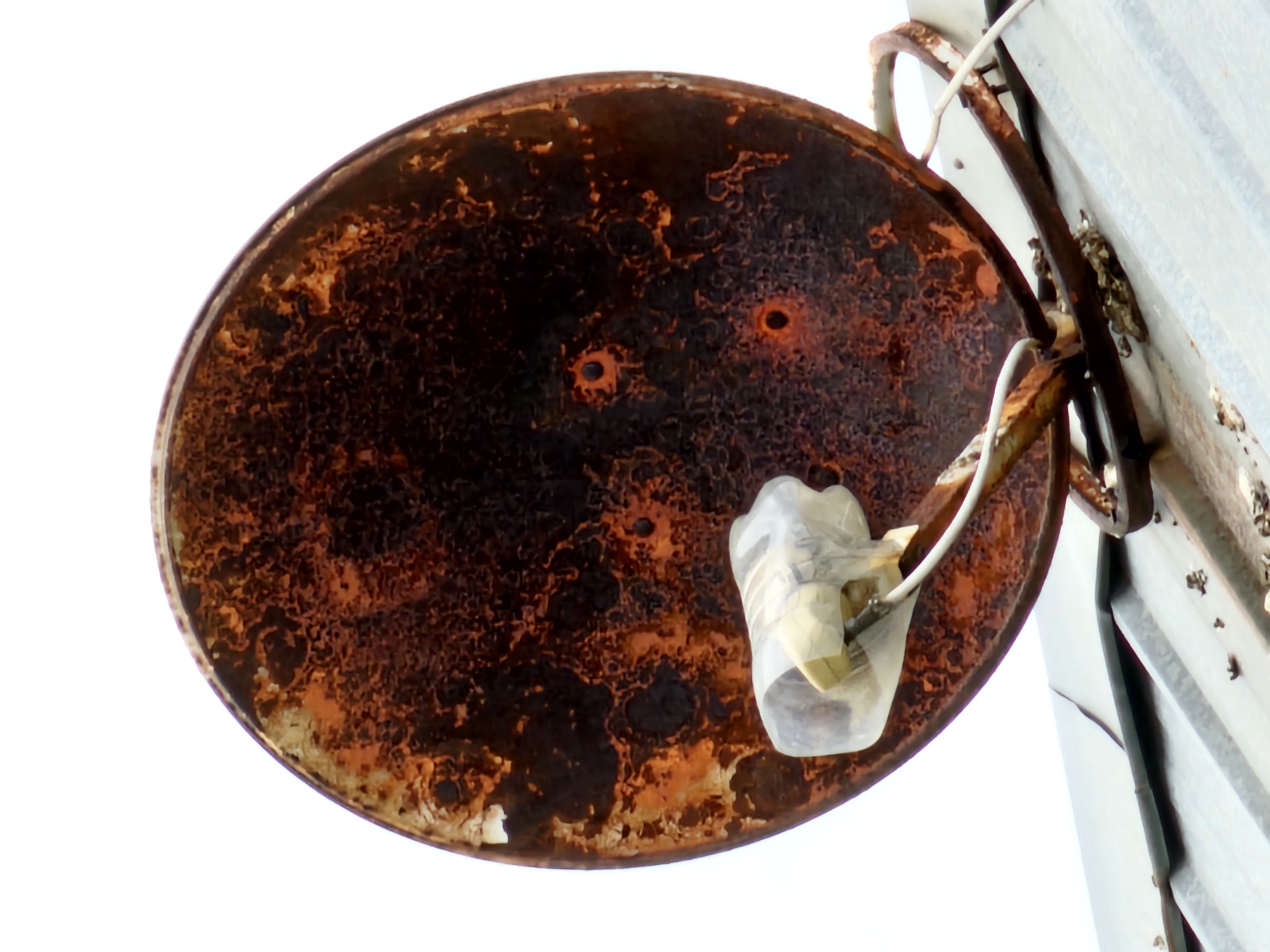 This image depicts a close-up, low-angle view of a rusty satellite dish mounted on a corrugated metal roof. The dish is heavily corroded, with large patches of rust and peeling paint, revealing a darker metal underneath. A white cable runs from the dish, and a transparent plastic wrap with tape is visible covering the receiver at the front of the dish. The background is a stark white, likely representing an overcast sky. The roof appears weathered and possibly in need of repair, with some debris scattered on its surface. The scene is devoid of human presence. The location is identified as Özgön, Kyrgyzstan.