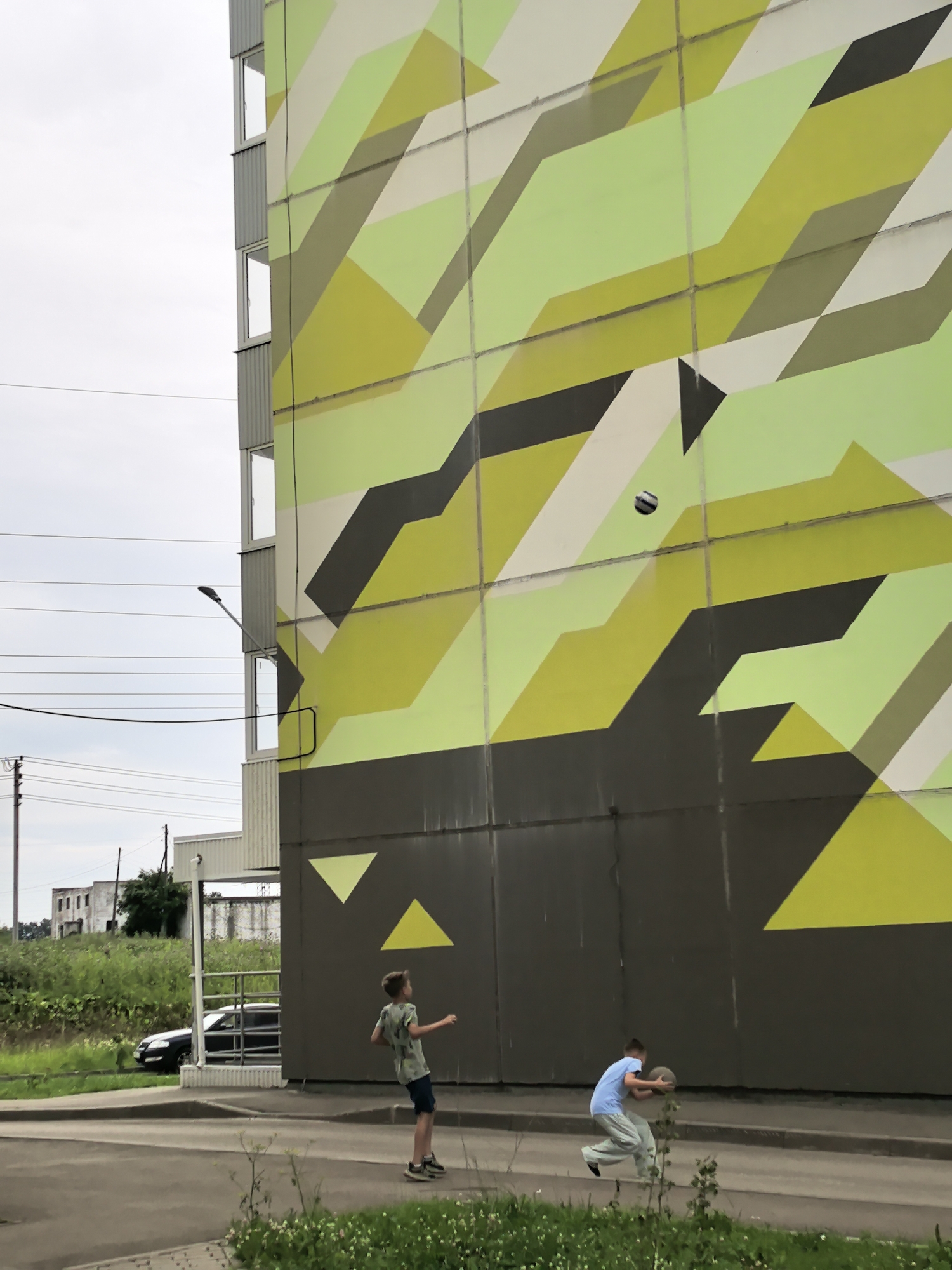 Two young boys are playing with a ball in front of a building with a large, abstract mural on its side. The mural features geometric shapes in shades of green, yellow, and grey. The boy on the left, wearing a t-shirt and shorts, is in mid-throw with the ball in the air. The boy on the right, wearing a blue t-shirt and light-colored pants, is bending down and appears to be catching or preparing to catch the ball. The scene is set outdoors on a paved area, with a sidewalk and some grassy patches visible. In the background, there are power lines, a street lamp, a parked car, and some buildings. The sky is overcast. The location is identified as Фролы, Russia.