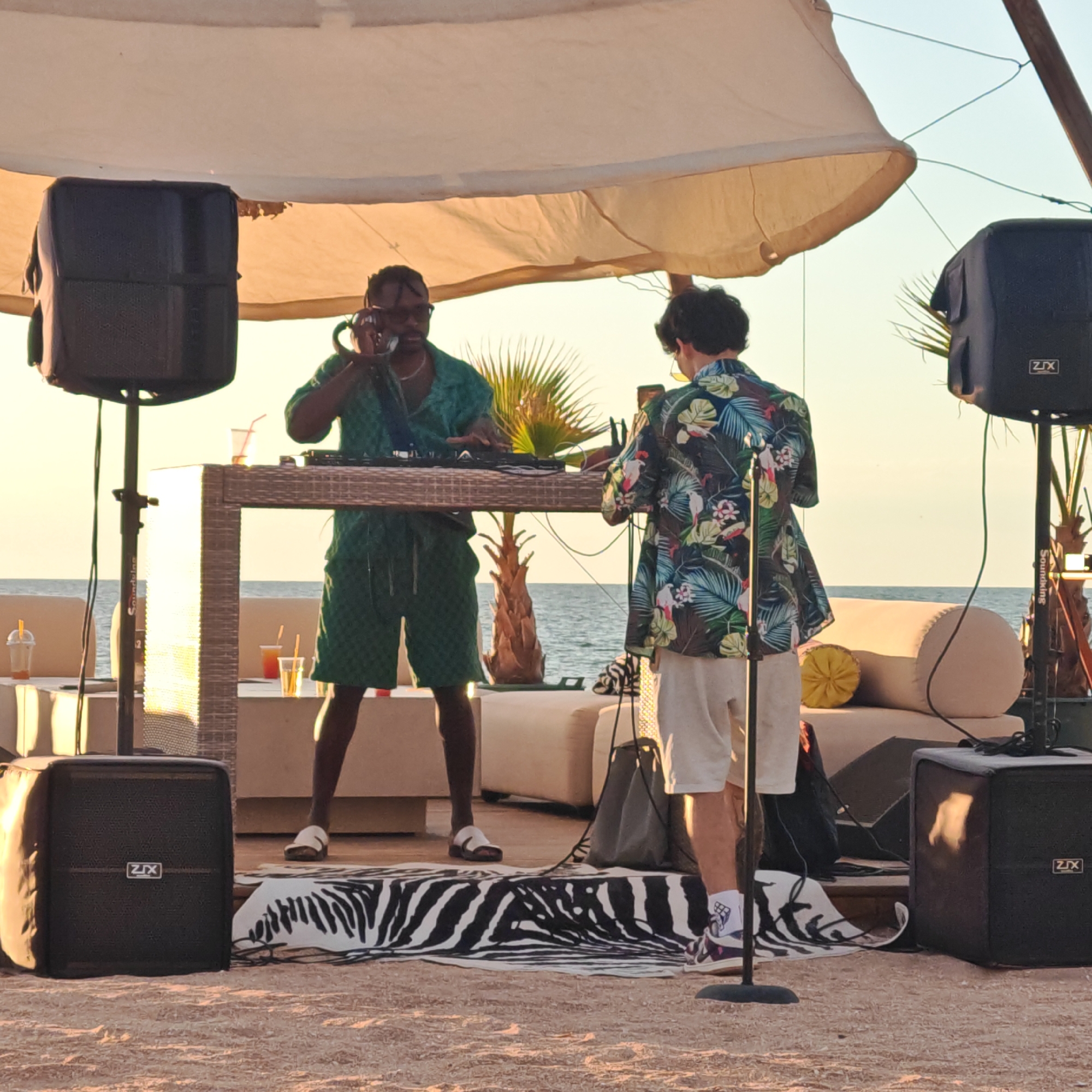 Two men are performing music outdoors near a body of water, possibly the sea. One man, wearing a green patterned short-sleeved shirt and green shorts, stands behind a DJ booth and appears to be operating equipment. He is holding a microphone to his ear. The other man, wearing a colorful tropical-patterned short-sleeved shirt and white shorts, stands to the right and holds a microphone. There are large black speakers on stands to the left and right of the DJ booth. A tan, woven table is set up in front of the DJ booth, with drinks on it. Behind the table and DJ booth, there are lounge chairs and a palm tree. The ground is covered in sand. The sky is clear and light. The scene appears to be a beach or seaside entertainment venue.