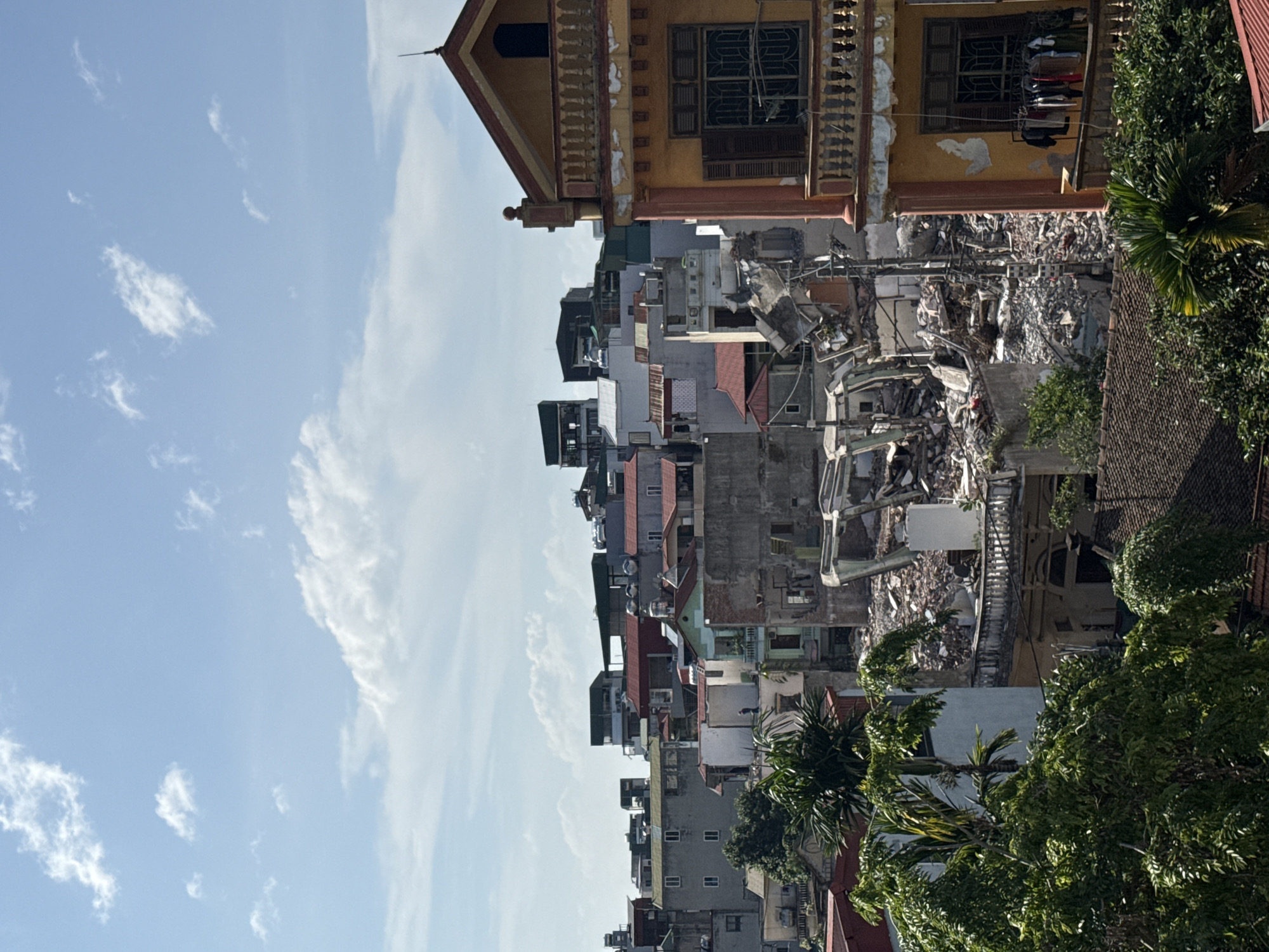 The image shows a vertical, eye-level view of a street scene in Hà Nội, Vietnam, featuring densely packed buildings ranging in height and architectural style. Some buildings are well-maintained, with visible details such as windows, balconies, and painted facades. In contrast, there is at least one building or structure that is partially collapsed or demolished. The rubble and debris are indicative of damage, possibly from construction, demolition, or some form of disaster. The sky is visible on the left side of the image, a bright blue with scattered white clouds. Green vegetation, including trees and plants, is visible on the right side, adding a natural element to the urban environment. Clothes hang on a drying rack from a balcony.