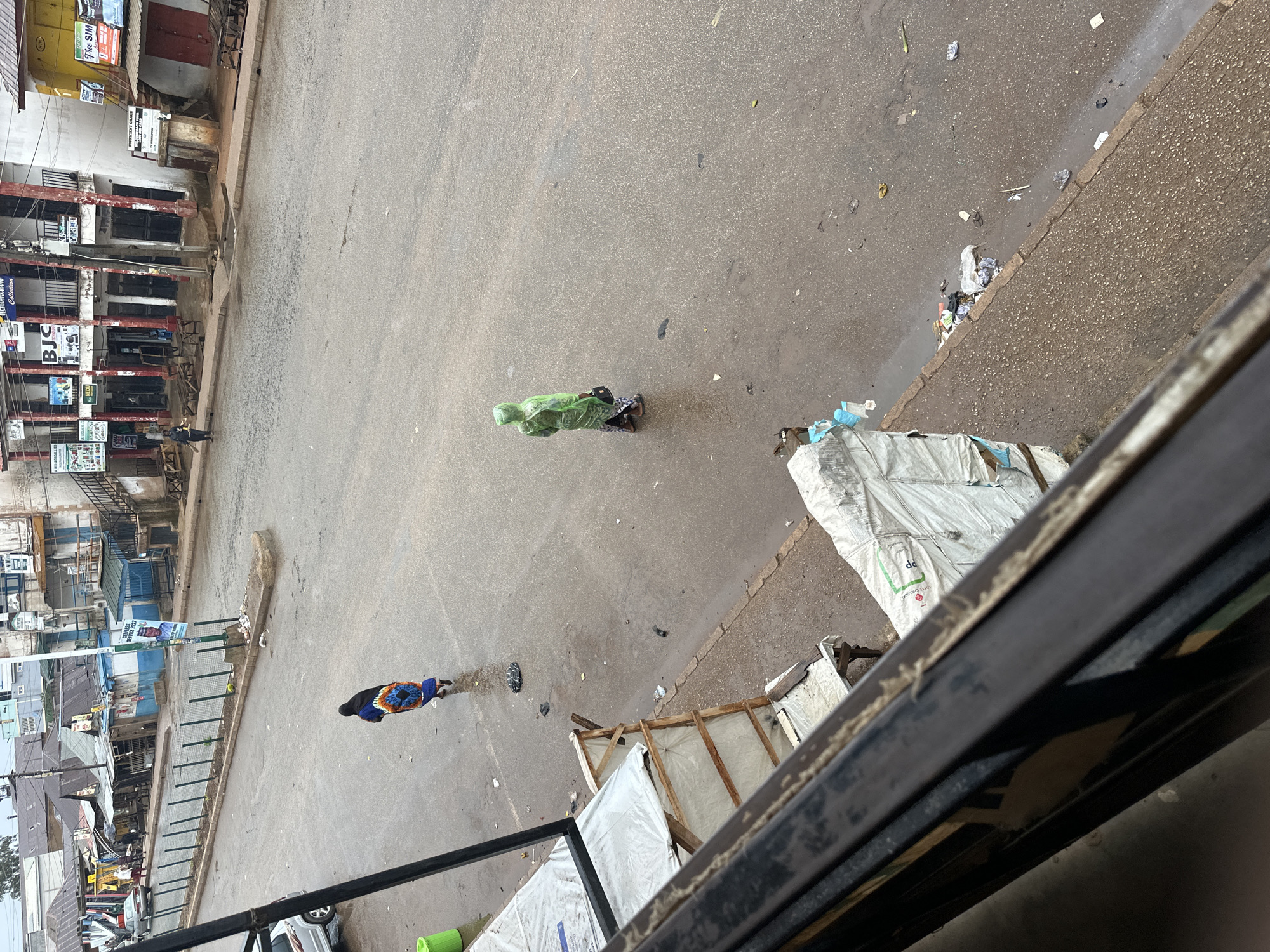 The image depicts a street scene in Bukuru, Nigeria, likely on a damp or rainy day given the wet asphalt and overcast sky. In the mid-ground, a person wearing a blue and orange patterned garment and a dark head covering walks away from the camera. Further to the right, another individual is seen from behind, wearing a green hooded raincoat and carrying a dark bag. The street is lined with buildings, some appearing to be shops or commercial establishments with visible signage. Several makeshift structures or market stalls covered with white tarps are present along the sidewalk on the left side of the image. Litter is scattered on the road and sidewalk, including plastic bags and paper debris. A silver car is parked on the left side of the street, and a green trash bin is visible near it. The overall impression is of a typical urban environment, possibly a market area, during a period of low activity.