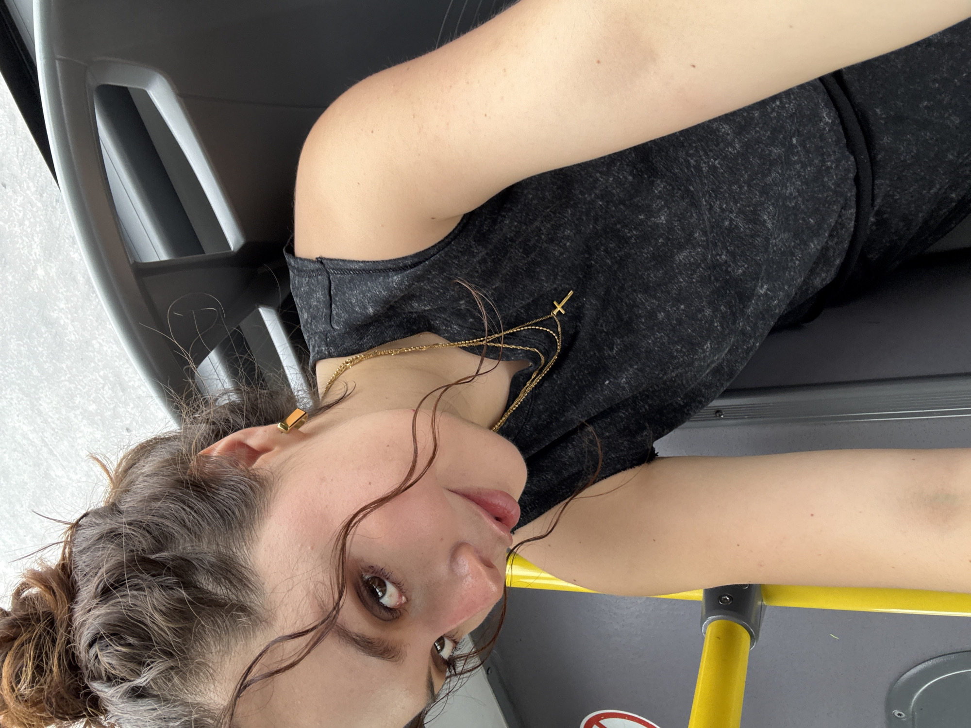 The image captures a young woman from a sideways perspective, appearing to be lying down or leaning inside what looks like a public transport vehicle, possibly in Apodaca, Mexico. She is the central subject, looking directly at the camera with a neutral expression.
She has medium-brown, curly hair, pulled back into a bun with several loose, damp-looking strands framing her face. She wears a dark grey or charcoal-colored sleeveless top. Around her neck are multiple thin golden necklaces, one of which features a small cross pendant. She also has a golden ear cuff visible on her left ear. Her skin tone is light.
The setting consists of grey plastic seating and a bright yellow metal handrail, common features in buses or trains. Below the handrail, a dark surface shows a partially visible white circular sticker with red, unreadable text. The lighting appears to be bright and even, suggesting daytime conditions. Some of her curly hair strands are visible on the grey surface of the seat near her arm. No specific weather conditions are discernible, but the indoor setting implies protection from the elements.