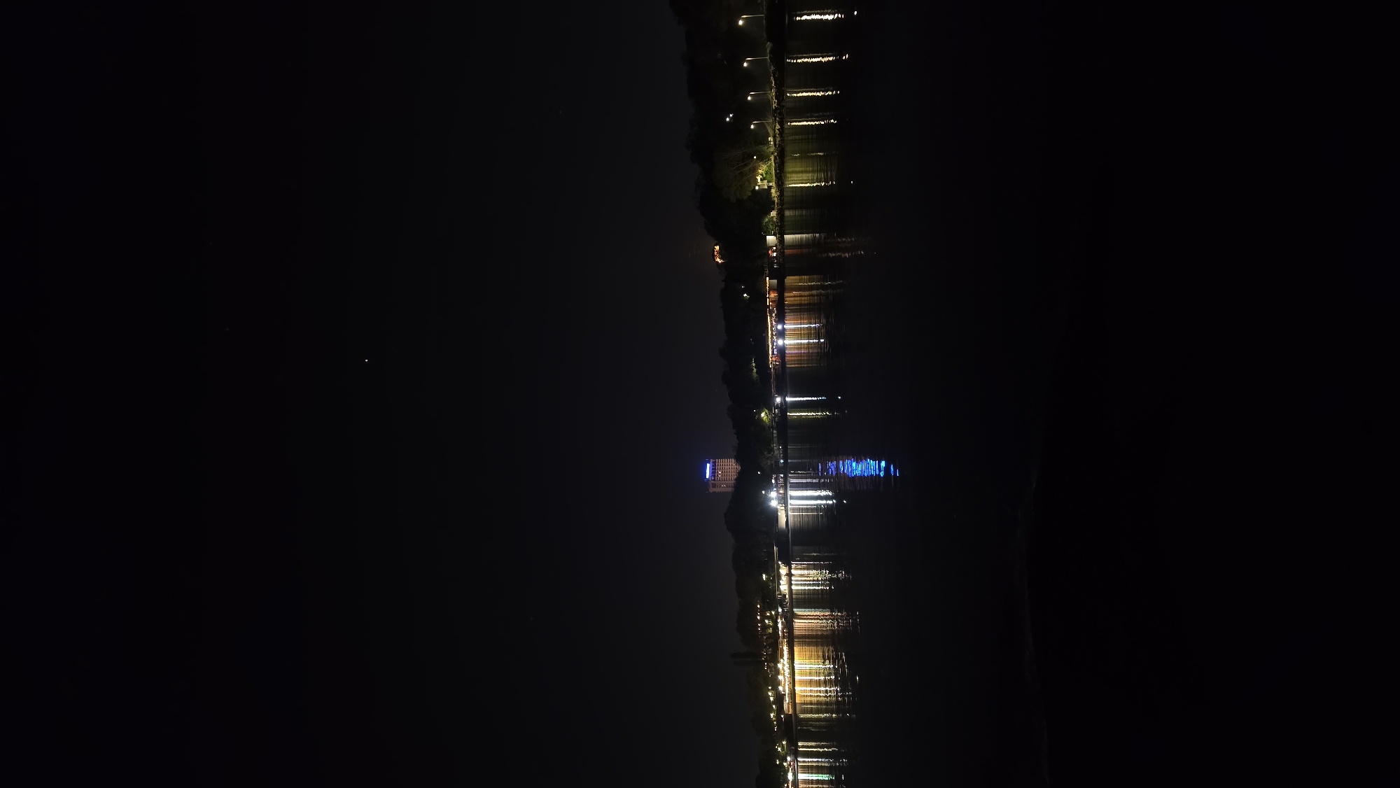 This image depicts the city of Varna, Bulgaria at night. The location appears to be along a waterfront area, potentially near a park or promenade, as evidenced by the presence of trees and lights along the edge. There is a body of water (possibly a lake or bay) reflecting the lights from the city. One building stands out due to its height and blue illumination on its upper floors. The reflection of lights on the water indicates either slight movement on the water's surface or a long exposure photograph. The rest of the scene is dark with only a few stars faintly visible in the sky. No people are visible in the image.